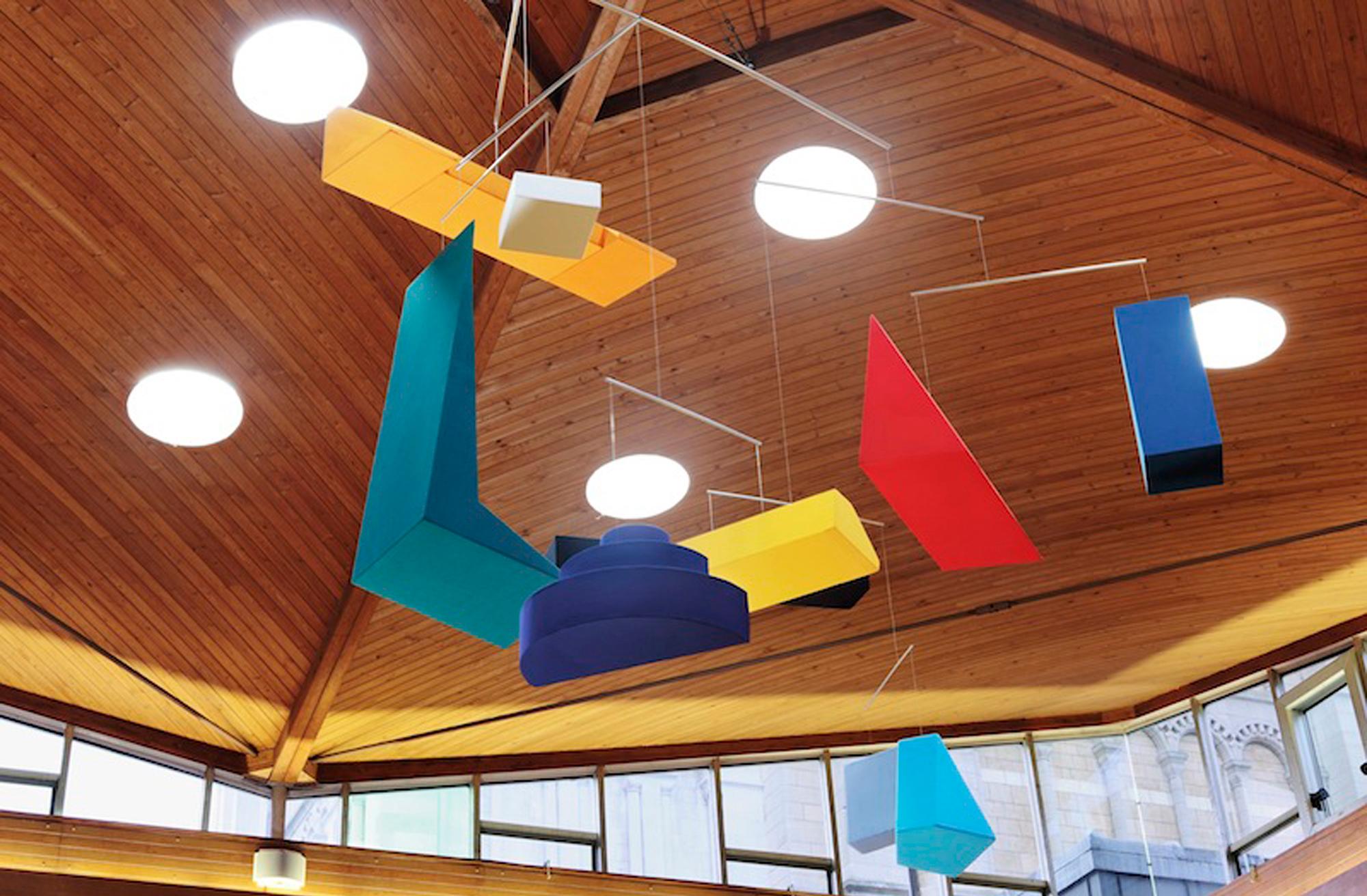 A vibrant abstract mobile with geometric shapes hangs from a wooden vaulted ceiling.