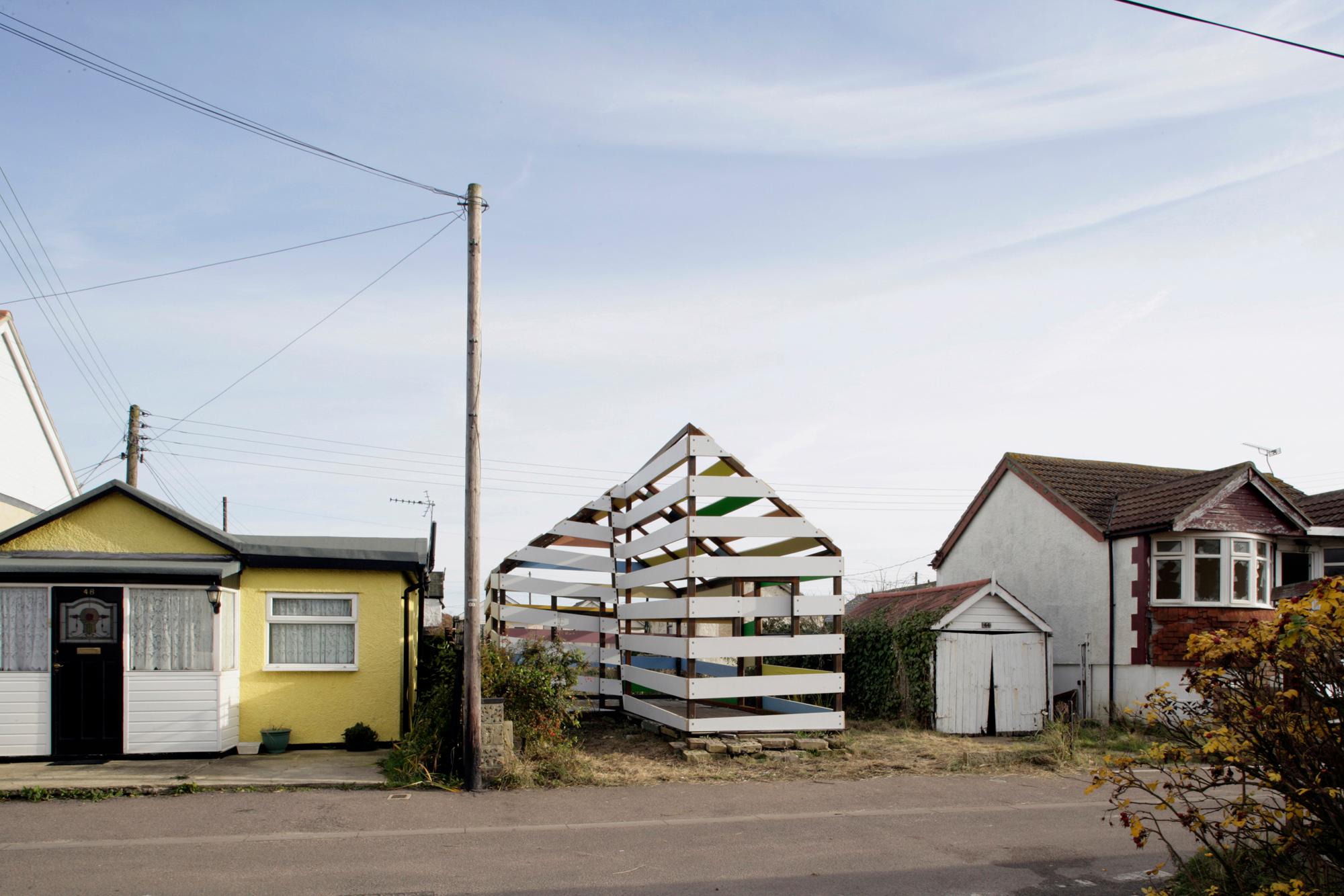 A street view showing a small yellow house, a utility pole, and an open, house-shaped frame made of white horizontal slats with colorful panels inside, next to a larger white and brick house.