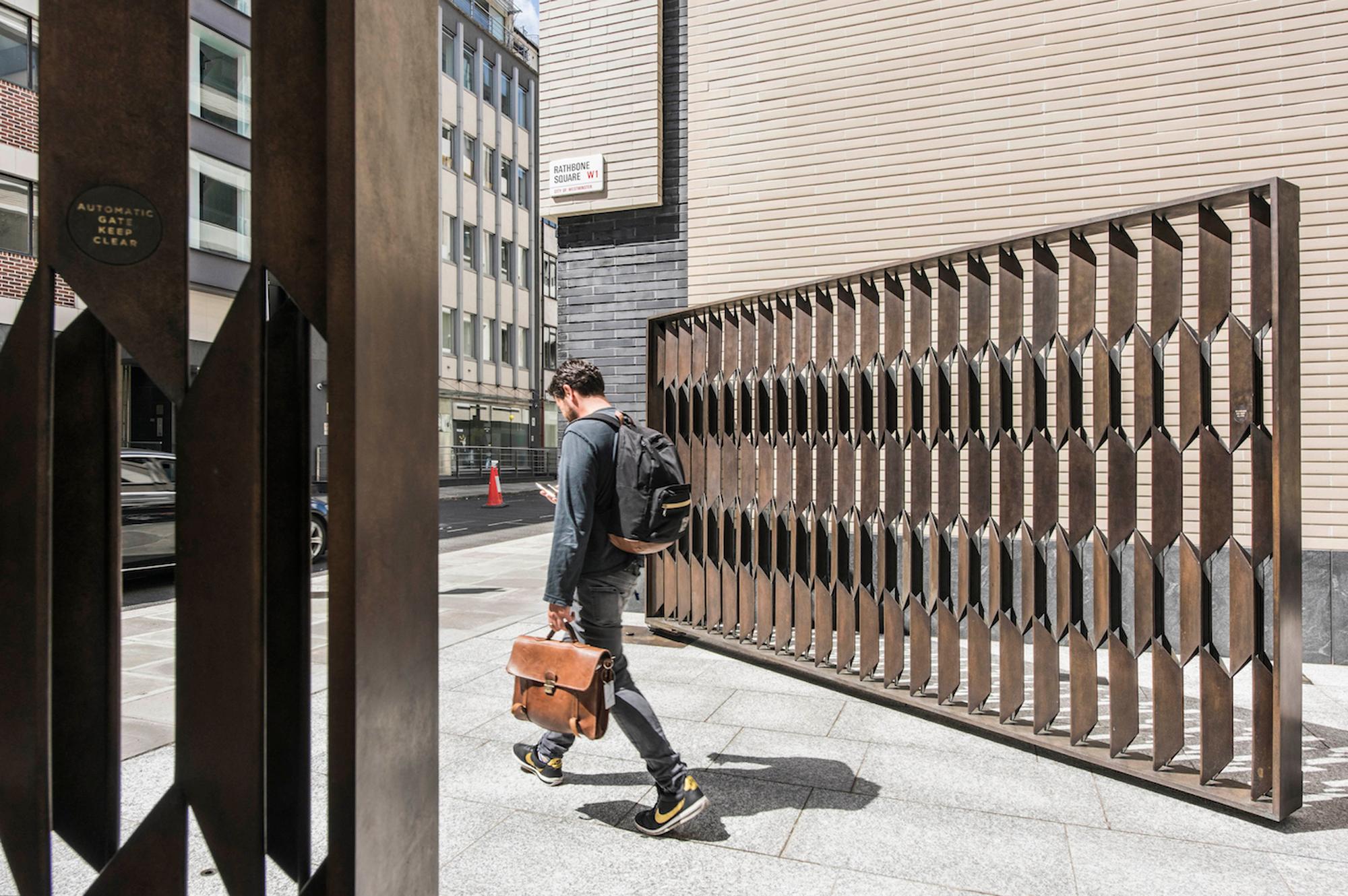 A man with a backpack and briefcase walks past modern metal structures, looking at his phone.