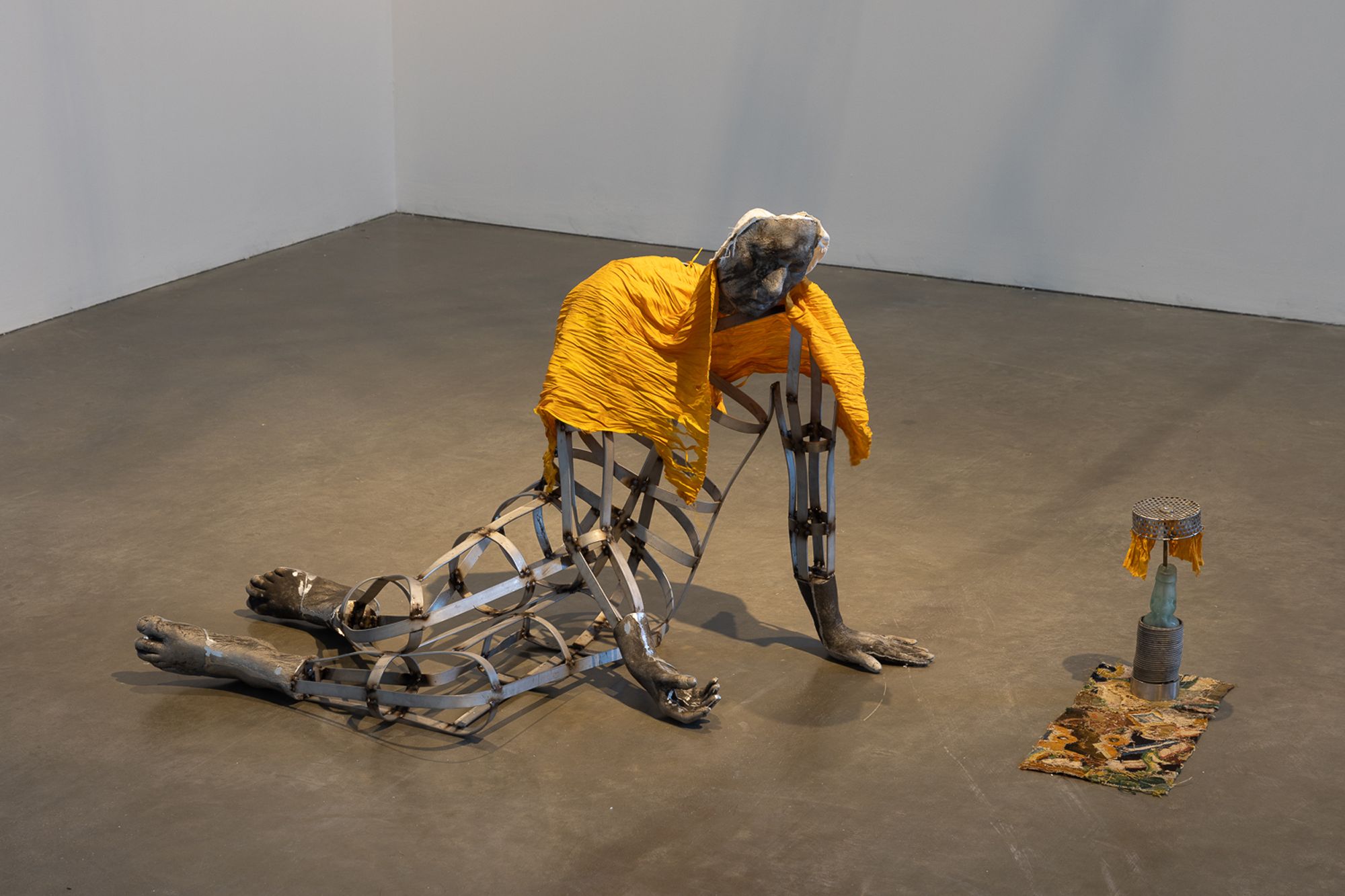 A metal sculpture of a hunched, cage-like human figure with solid legs, draped in yellow fabric. A smaller cylindrical sculpture is nearby on a concrete floor.