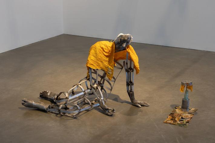 A metal sculpture of a hunched, cage-like human figure with solid legs, draped in yellow fabric. A smaller cylindrical sculpture is nearby on a concrete floor.