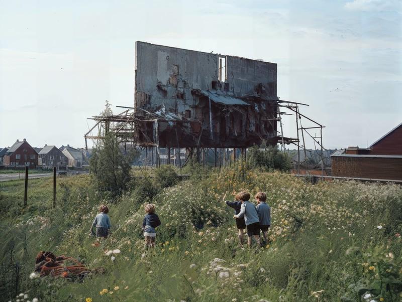 Several children play in a field of tall wildflowers with a large, ruined screen structure and houses in the background.