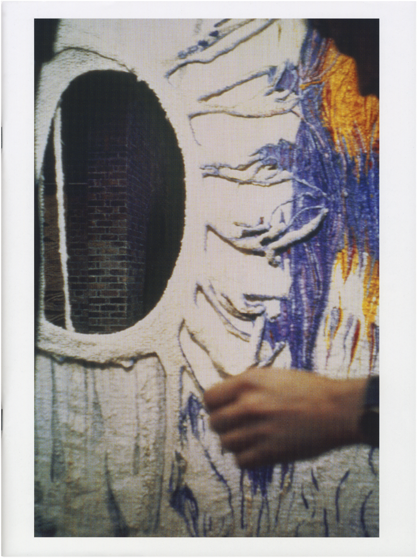 Textured textile art with an oval cutout revealing a brick wall, featuring blue and orange weaving and a hand touching the fabric.