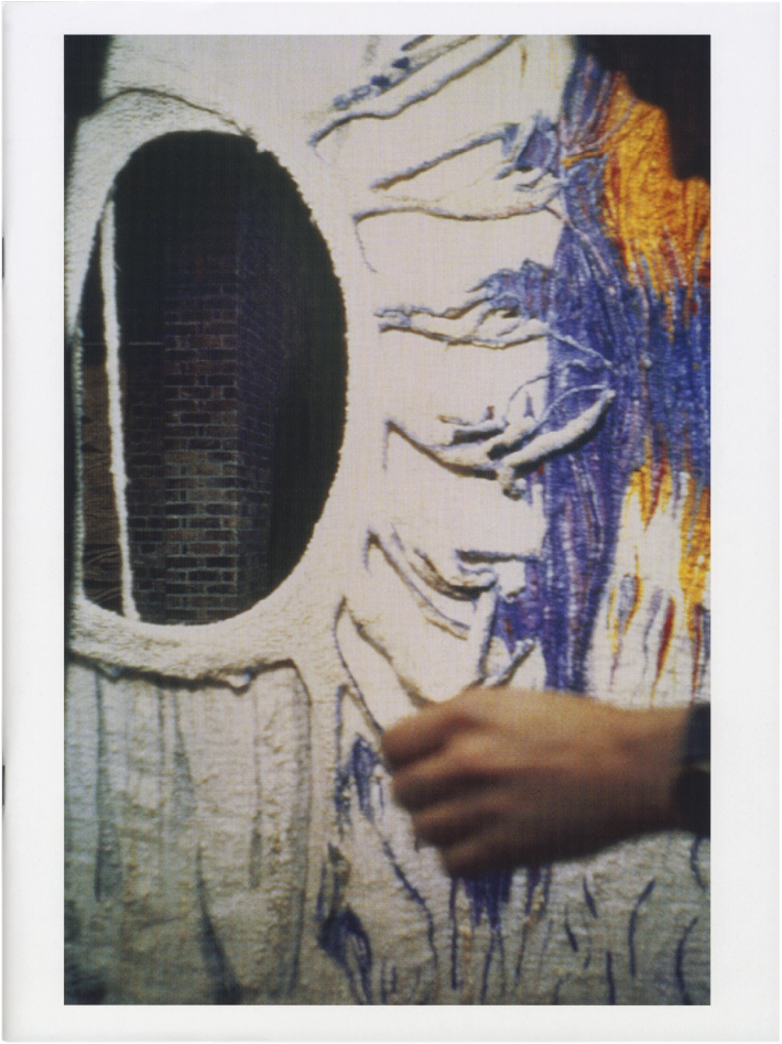 Textured textile art with an oval cutout revealing a brick wall, featuring blue and orange weaving and a hand touching the fabric.