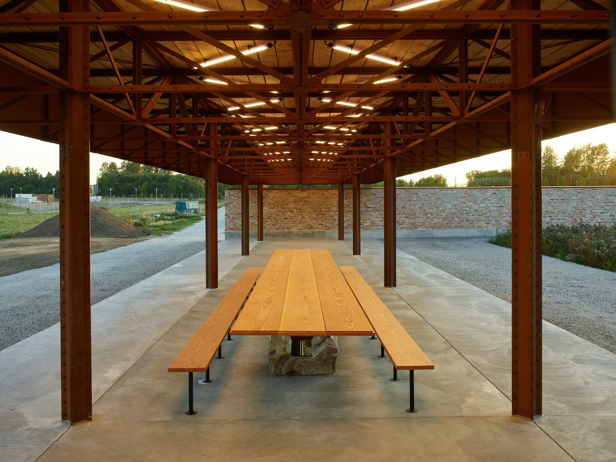 A long outdoor pavilion with a wooden roof, metal columns, and a large wooden picnic table with benches on a concrete floor.