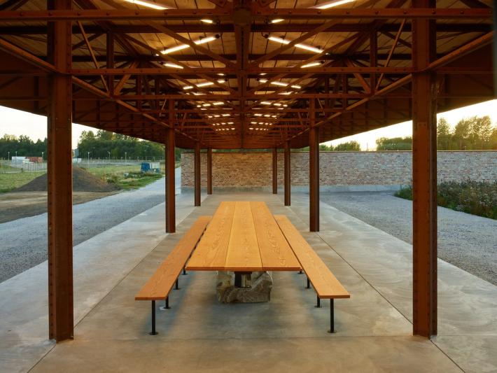 A long outdoor pavilion with a wooden roof, metal columns, and a large wooden picnic table with benches on a concrete floor.