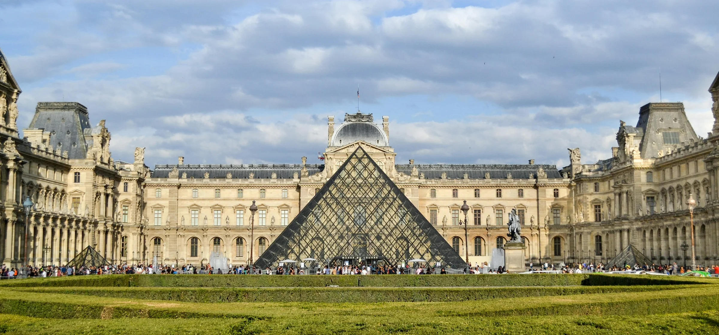 Piramida kaca dan bangunan Museum Louvre di Paris dengan langit biru berawan.