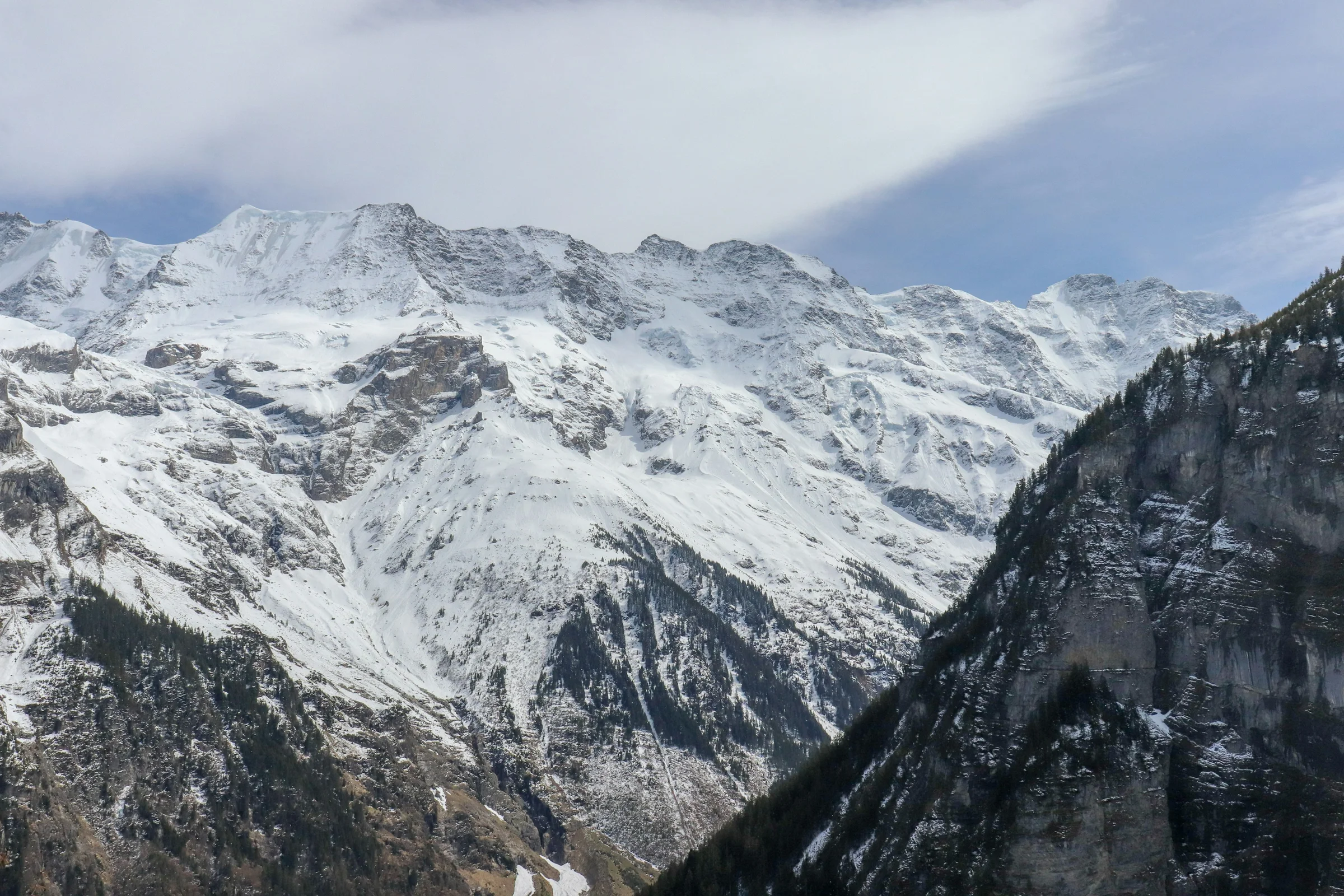 Lembah pegunungan Swiss dengan lereng dan puncak Alpen berselimut salju di bawah langit berawan.