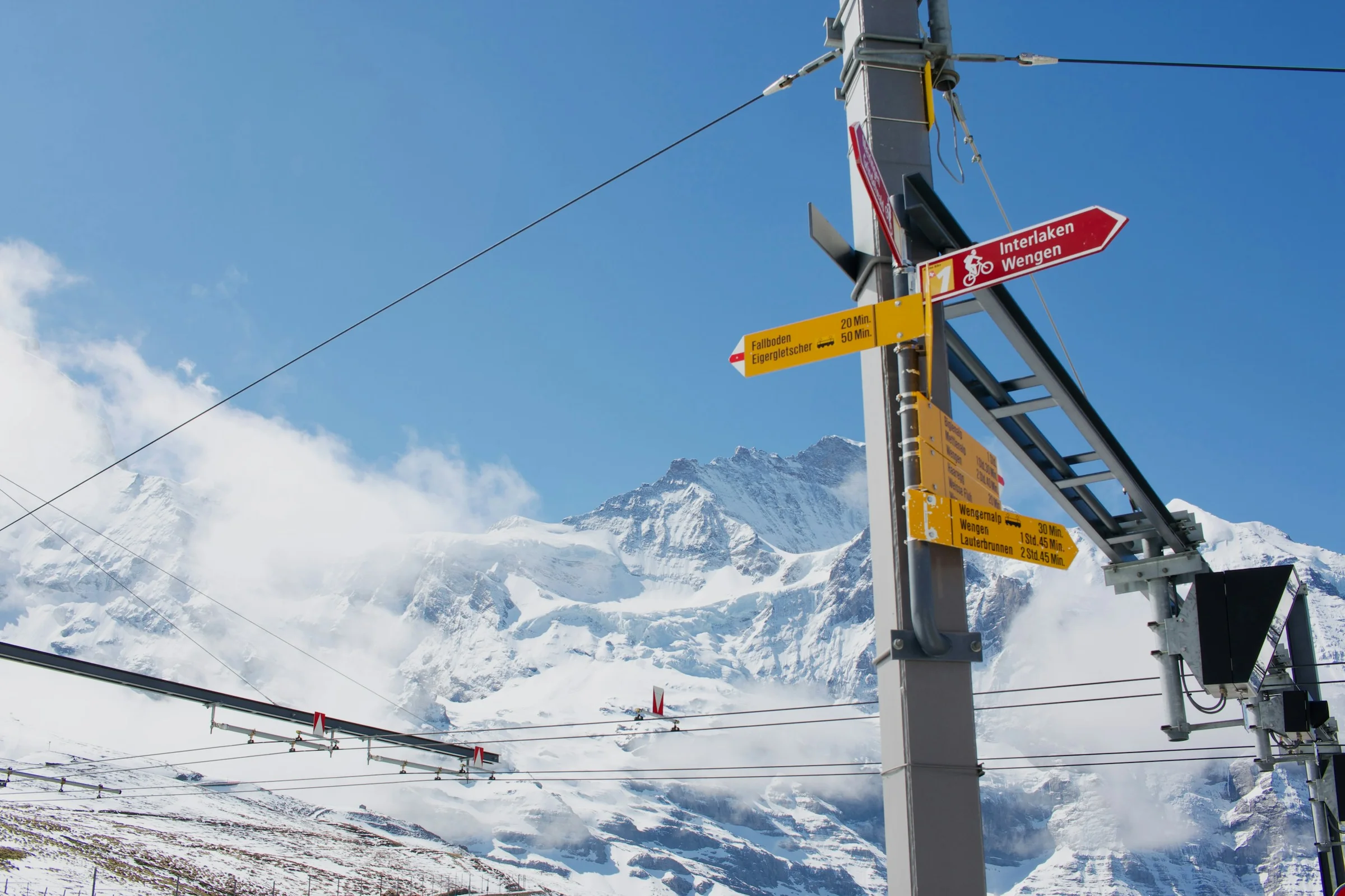 Papan petunjuk jalur pendakian dengan kabel gondola dan puncak Alpen bersalju di belakangnya.