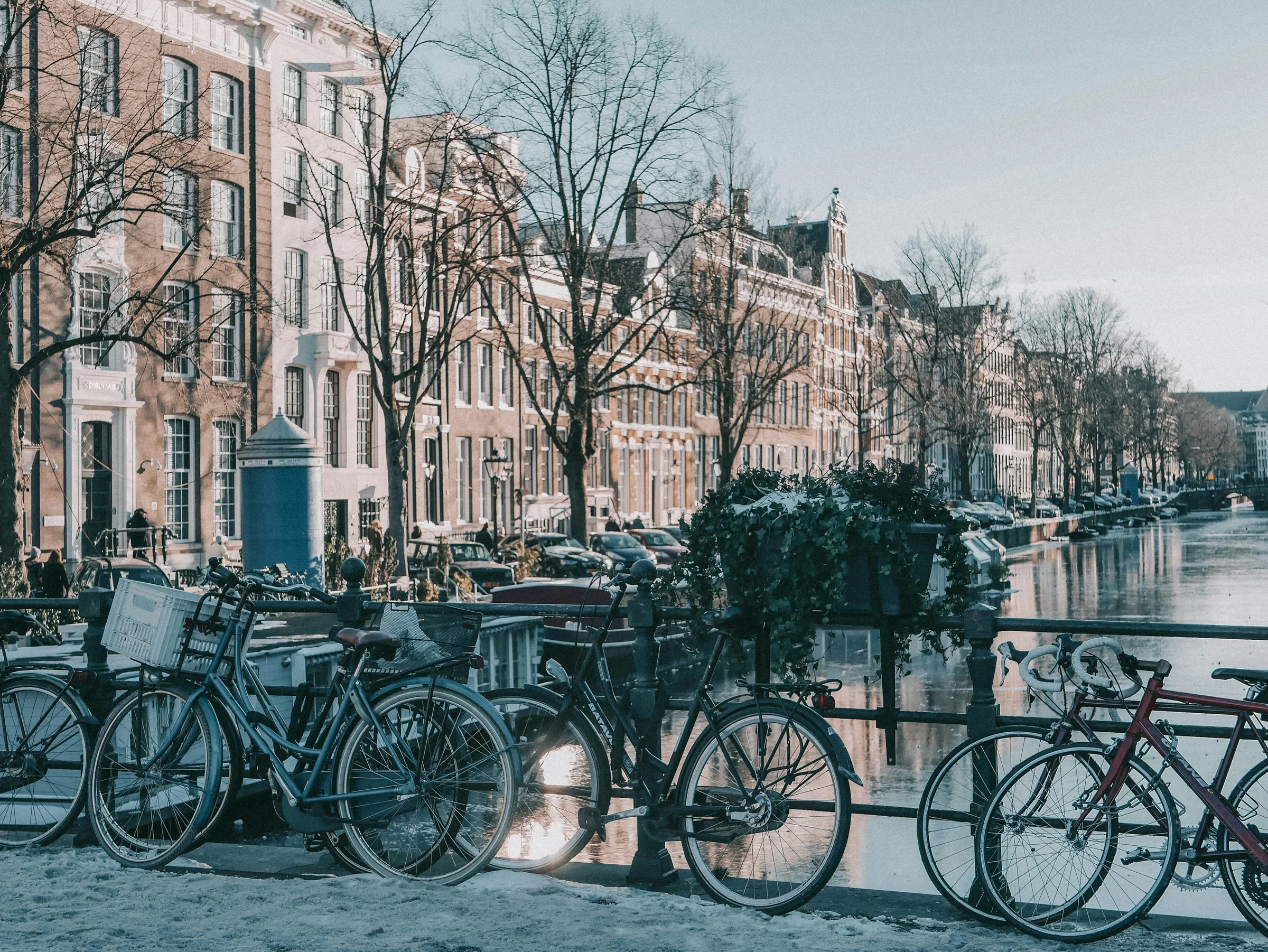 Kanal Amsterdam di musim dingin dengan pohon gundul, deretan rumah batu, dan sepeda bersandar di pagar tepi kanal.