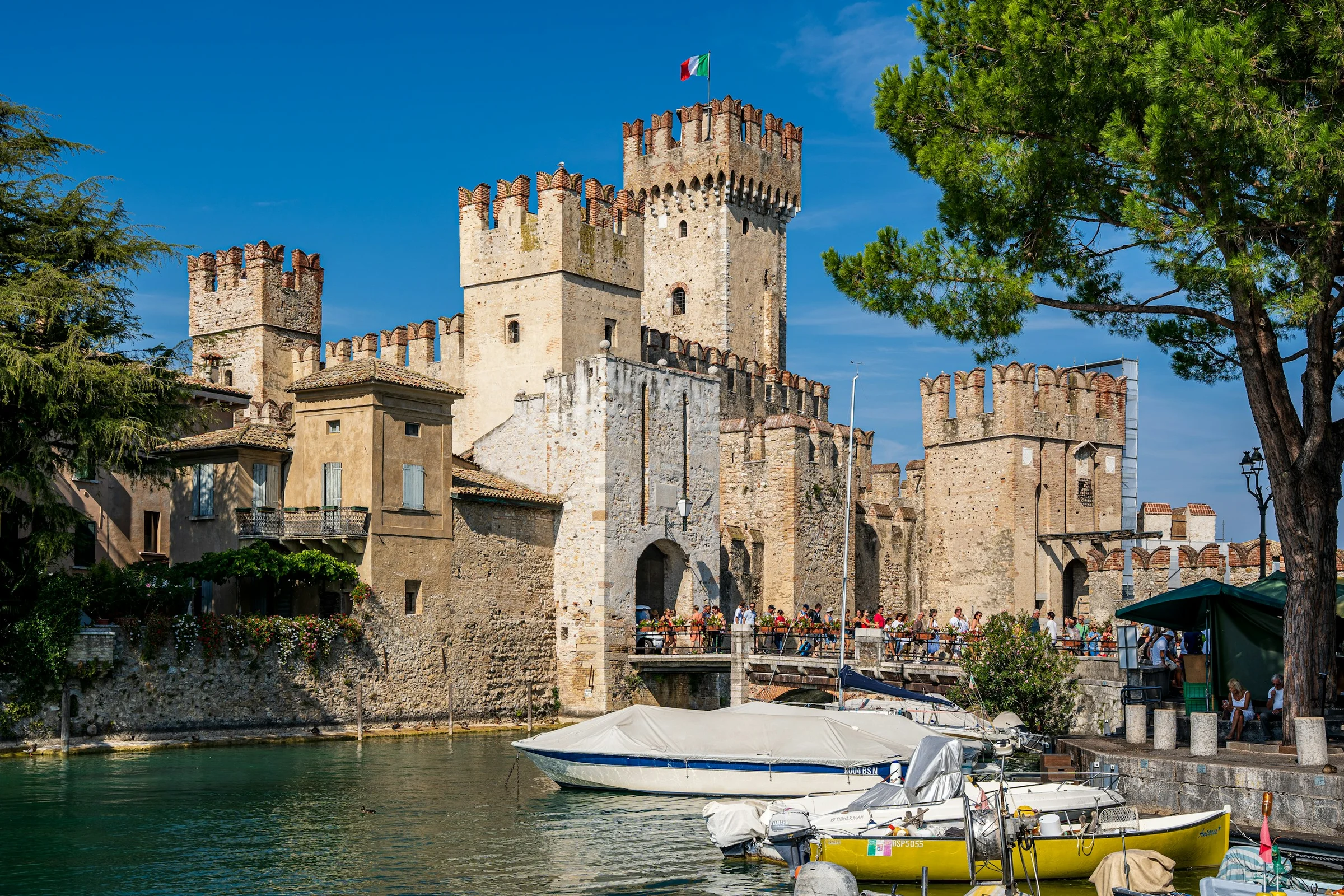 Kastil Scaligero Sirmione dengan pohon pinus dan perahu di Danau Garda di bawah langit biru.