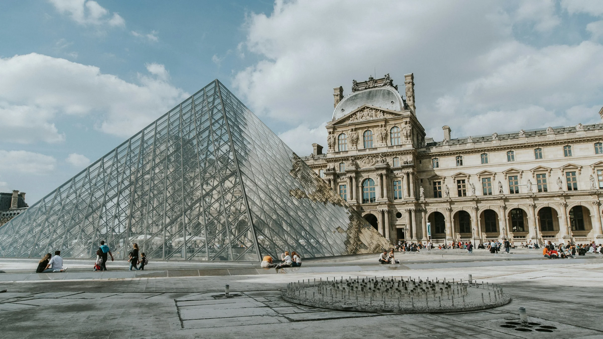 Piramida kaca dan bangunan Museum Louvre di bawah langit berawan, Paris.