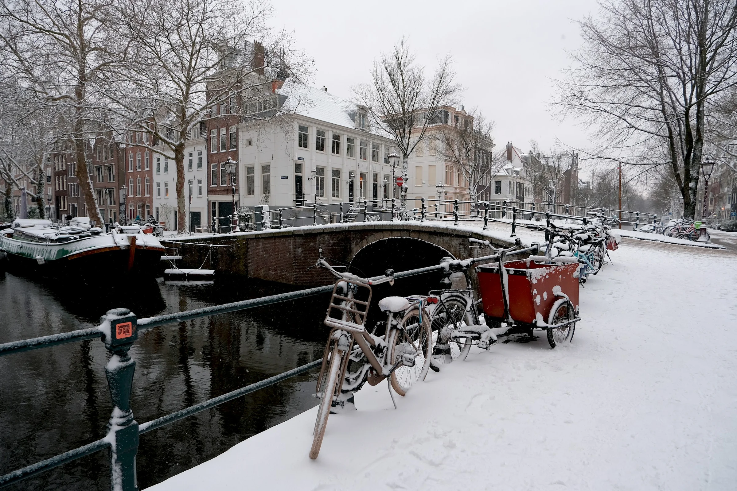 Jembatan kanal Amsterdam dengan sepeda terparkir dan salju menutupi jalan tepi kanal.