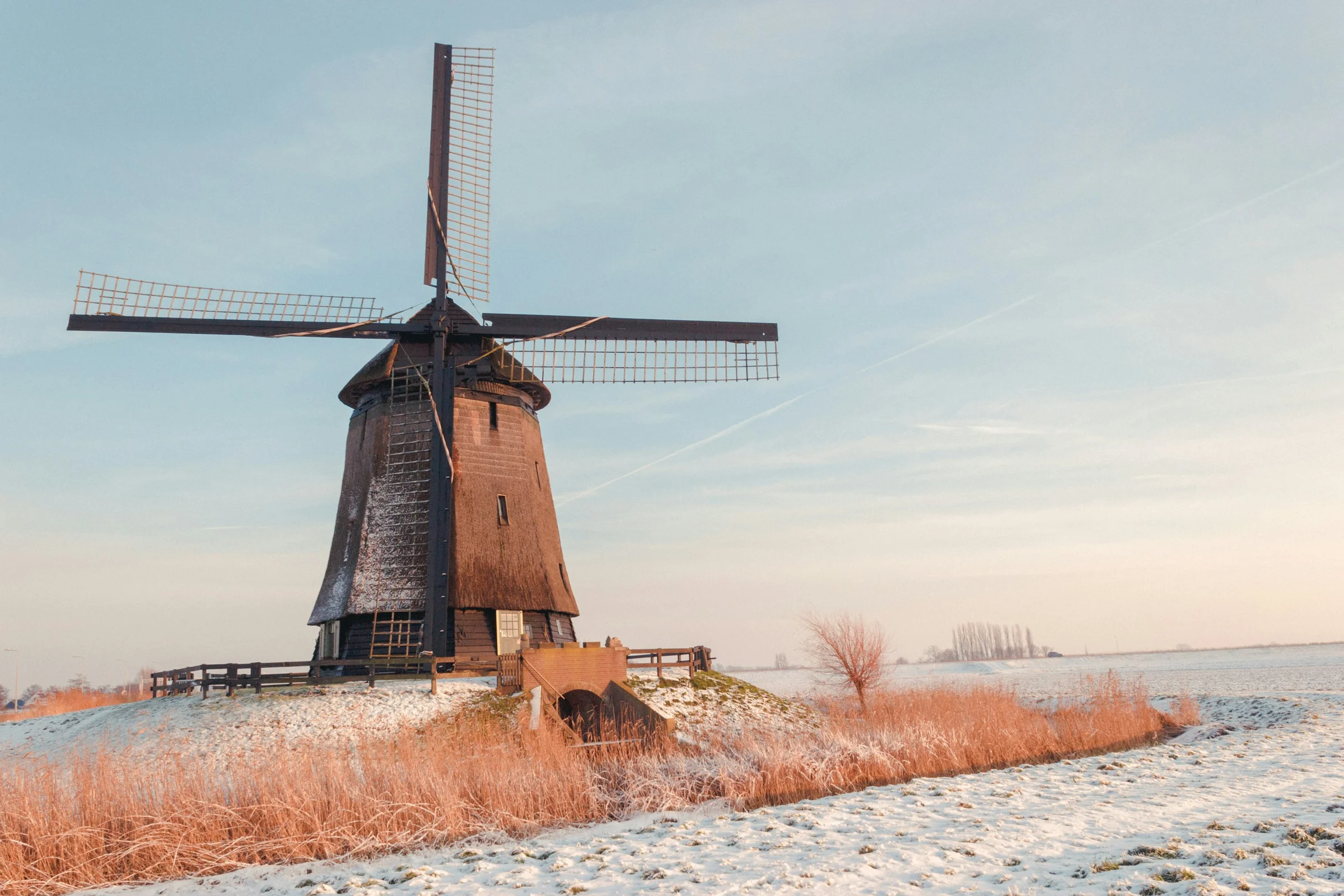 Kincir angin di padang polder Belanda yang bersalju dengan langit pucat berwarna lembut.