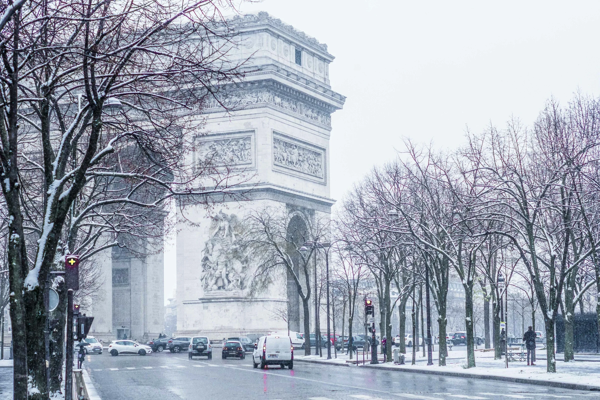 Arc de Triomphe di Paris dengan pohon gundul bersalju dan lalu lintas di sekeliling bundaran.