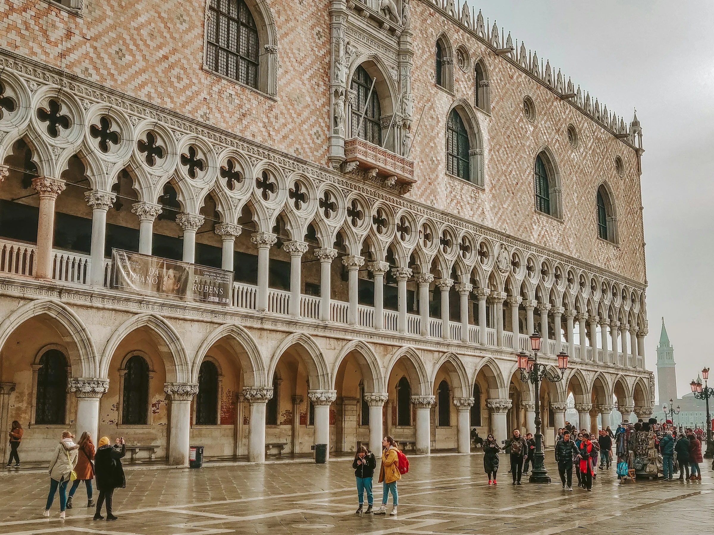 Fasad Doge's Palace di Venice di bawah langit mendung dengan wisatawan kecil di bawahnya.