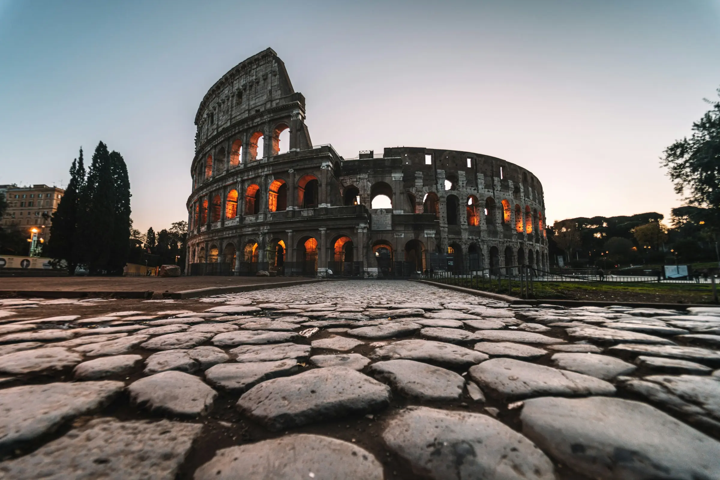 Colosseum Roma menyala keemasan saat senja dengan jalan berbatu kuno di depannya