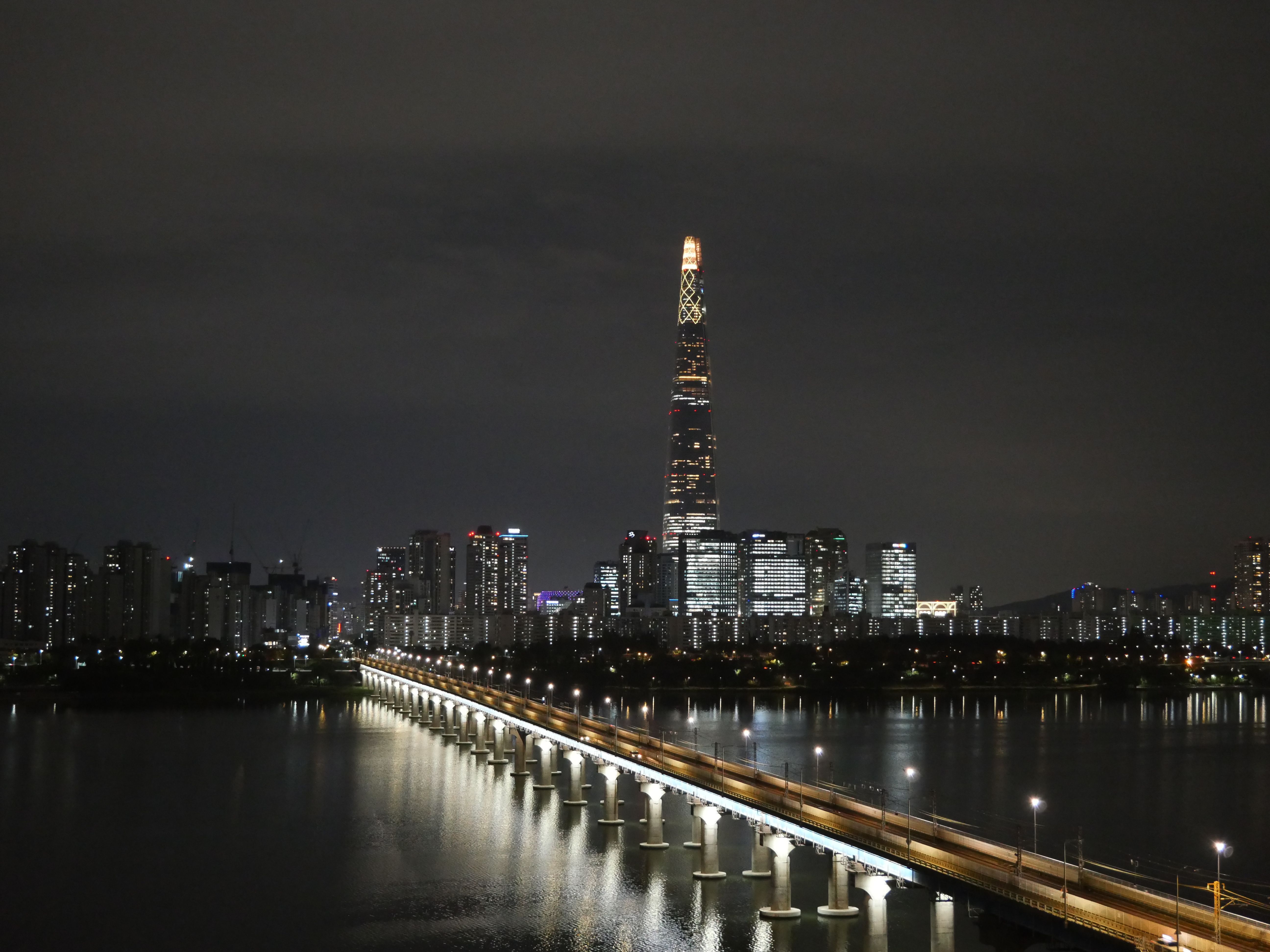 Lotte World Tower menyala malam hari di atas Sungai Han dengan jembatan lurus bercahaya