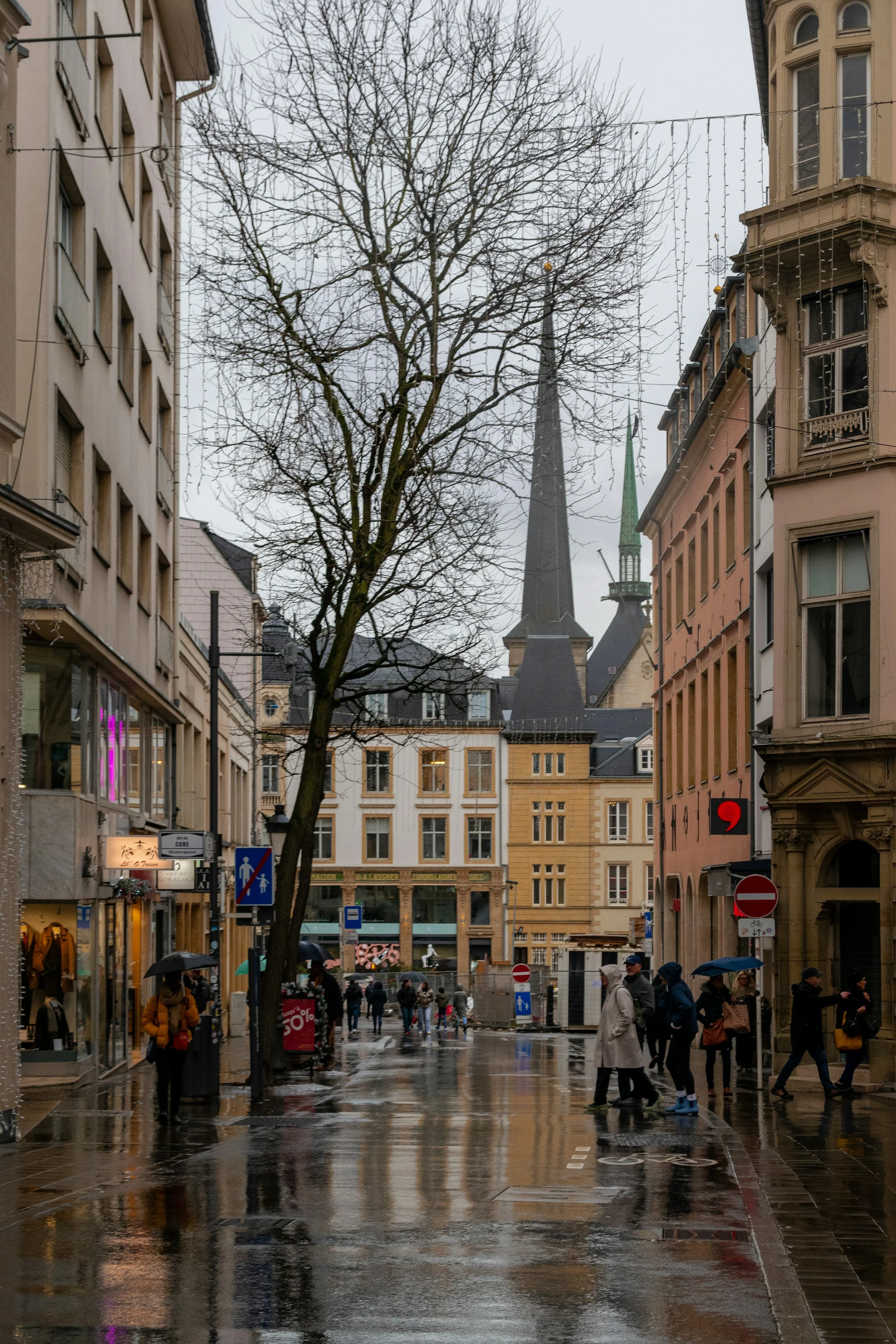 Jalan kota tua Luxembourg yang basah dengan menara Katedral Notre-Dame di ujung jalan dan pohon gundul.