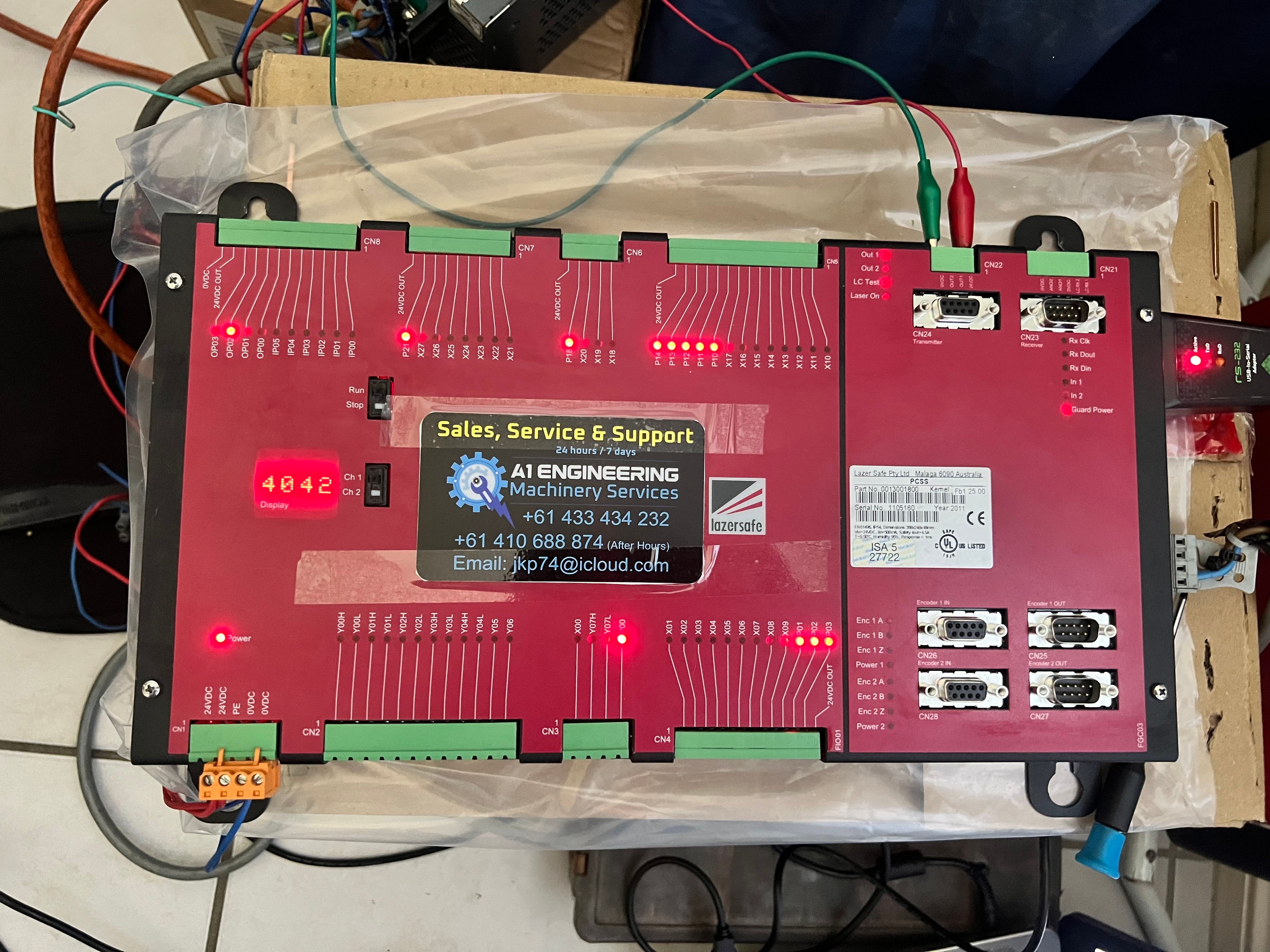 A red Lazer Safe PCSS safety controller board with lit indicator lights, connectors and test leads laid out for bench testing.