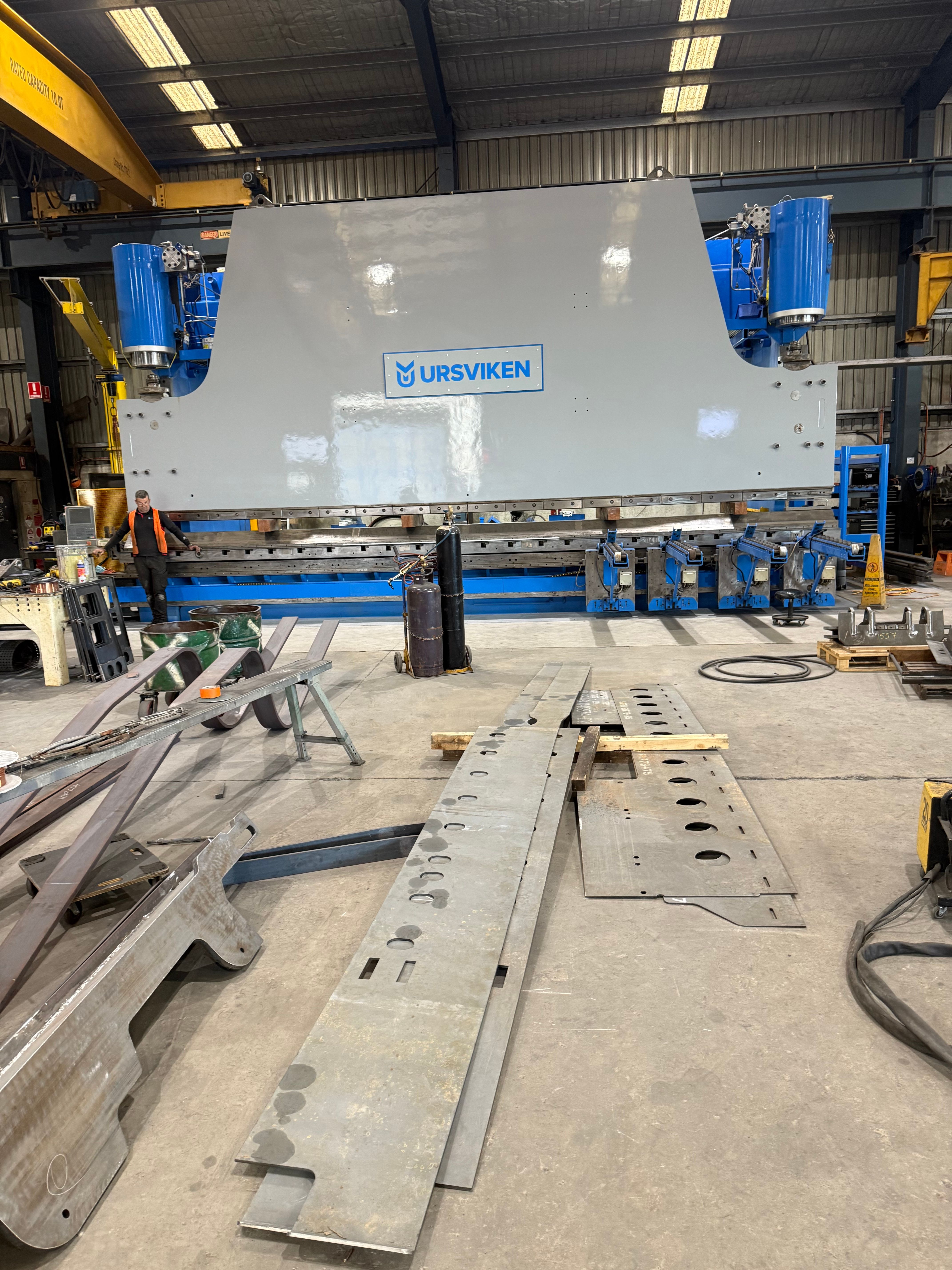 A large repainted Ursviken press brake with blue hydraulic cylinders and backgauge, with cut steel plate components laid out on the workshop floor.