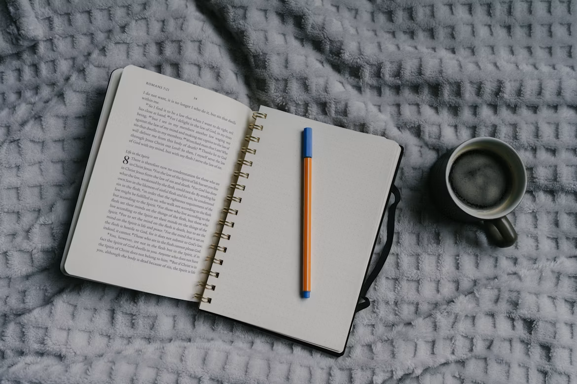An open Bible beside a notebook, pen, and cup of coffee