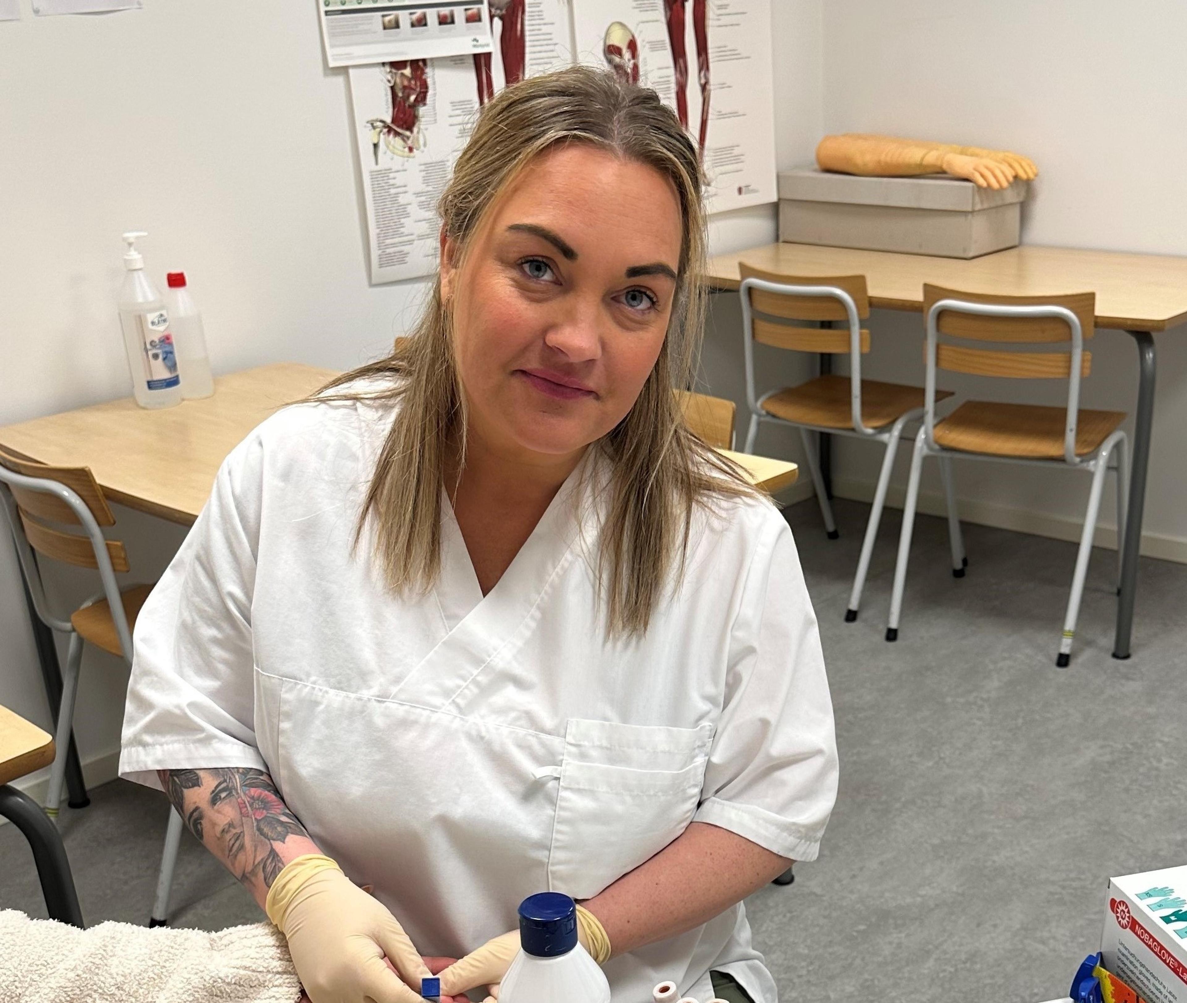 Ung dame i hvit frakk sitter i Folkeuniversitetets elevklinikk og øver på å ta blodprøver.