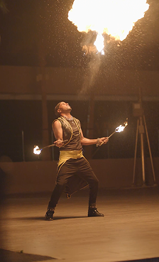 Fire performer holding flaming torches beneath a plume of fire.