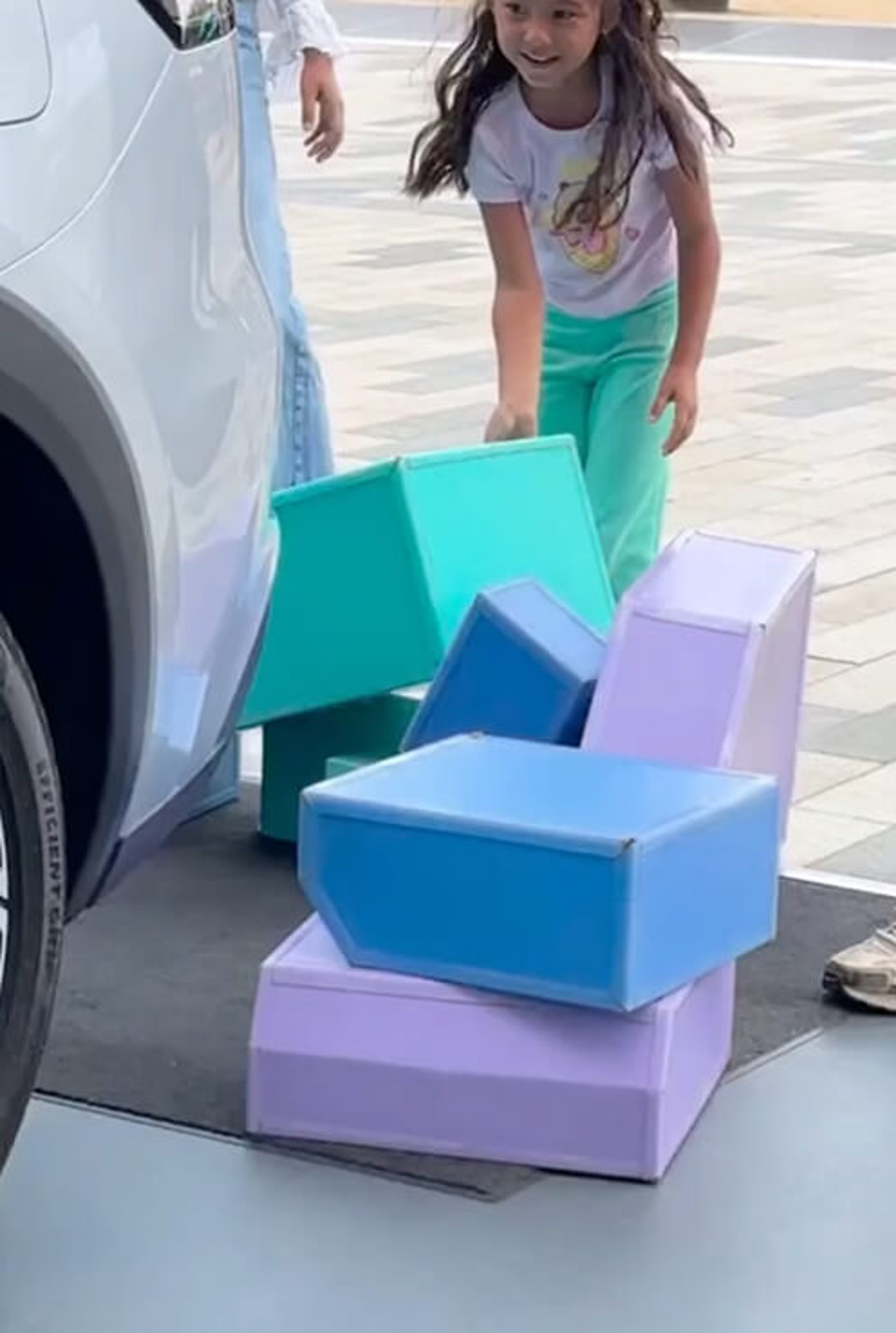 A young girl playing with large colorful blocks on the ground next to a car