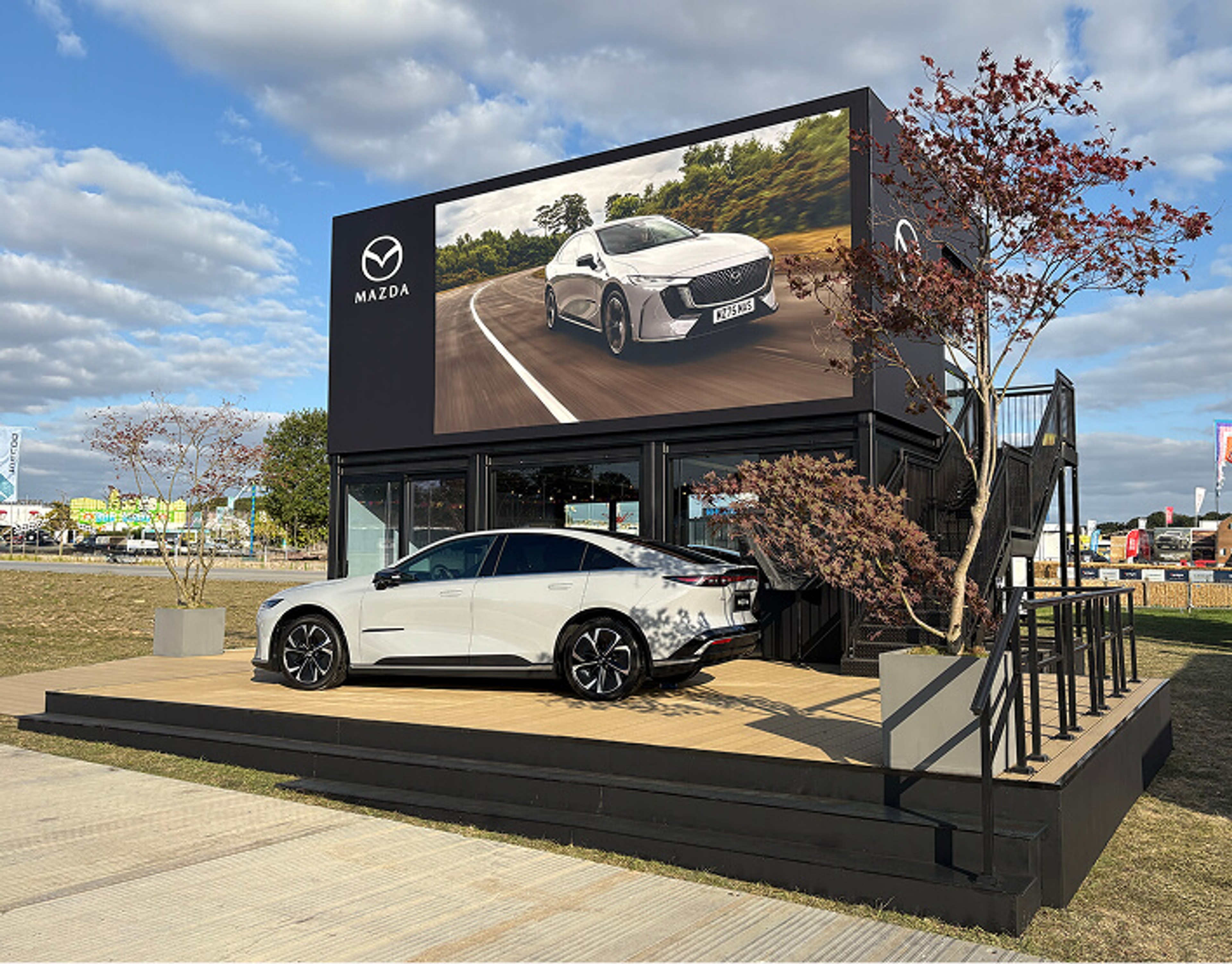 A white Mazda sedan parked on display in front of a black multi-story Mazda exhibition stand with a large outdoor LED screen
