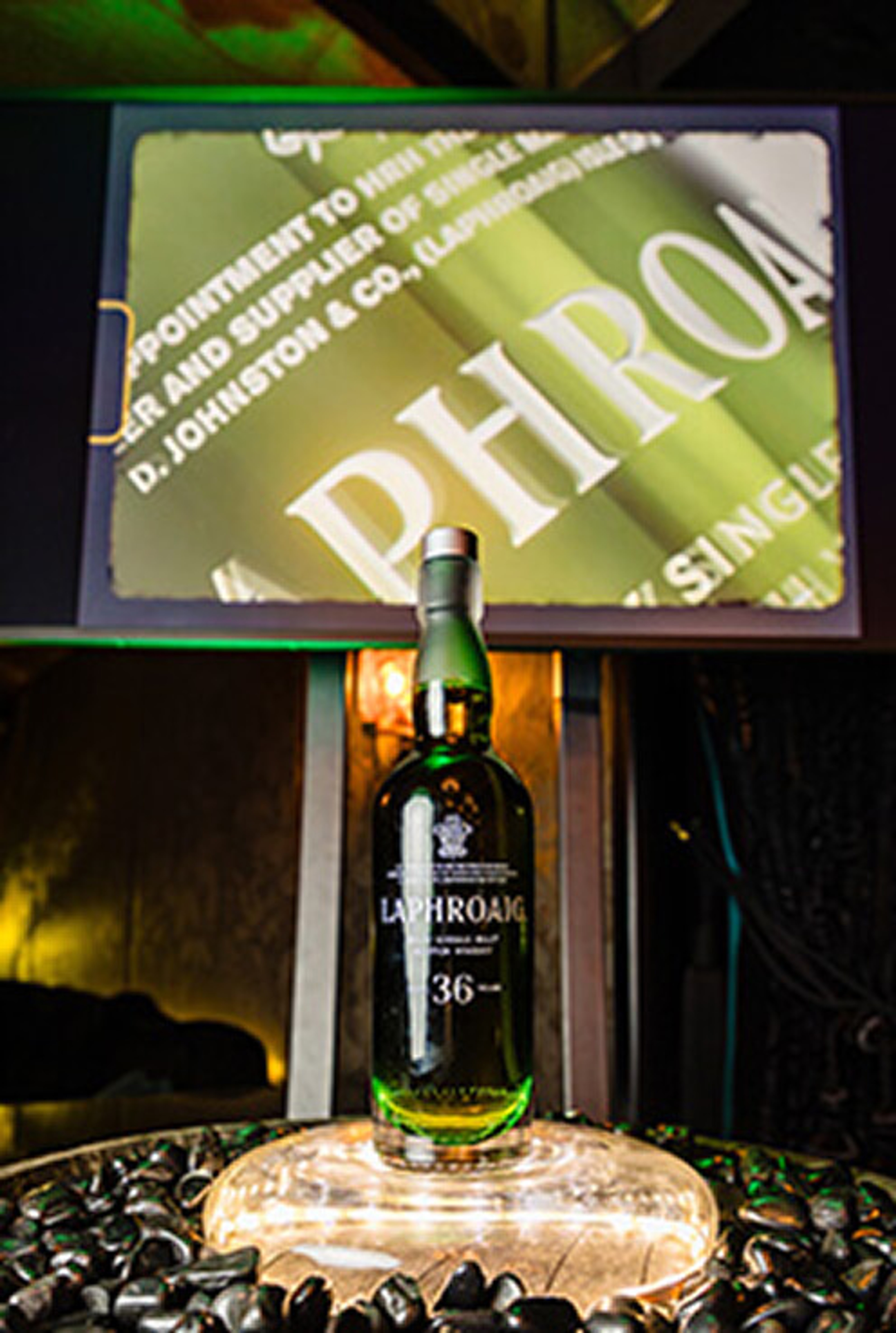 A green bottle of Laphroaig 36-Year-Old whisky standing on an illuminated base in front of a screen