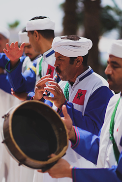 Moroccan musicians in traditional dress playing a drum and wind instrument.