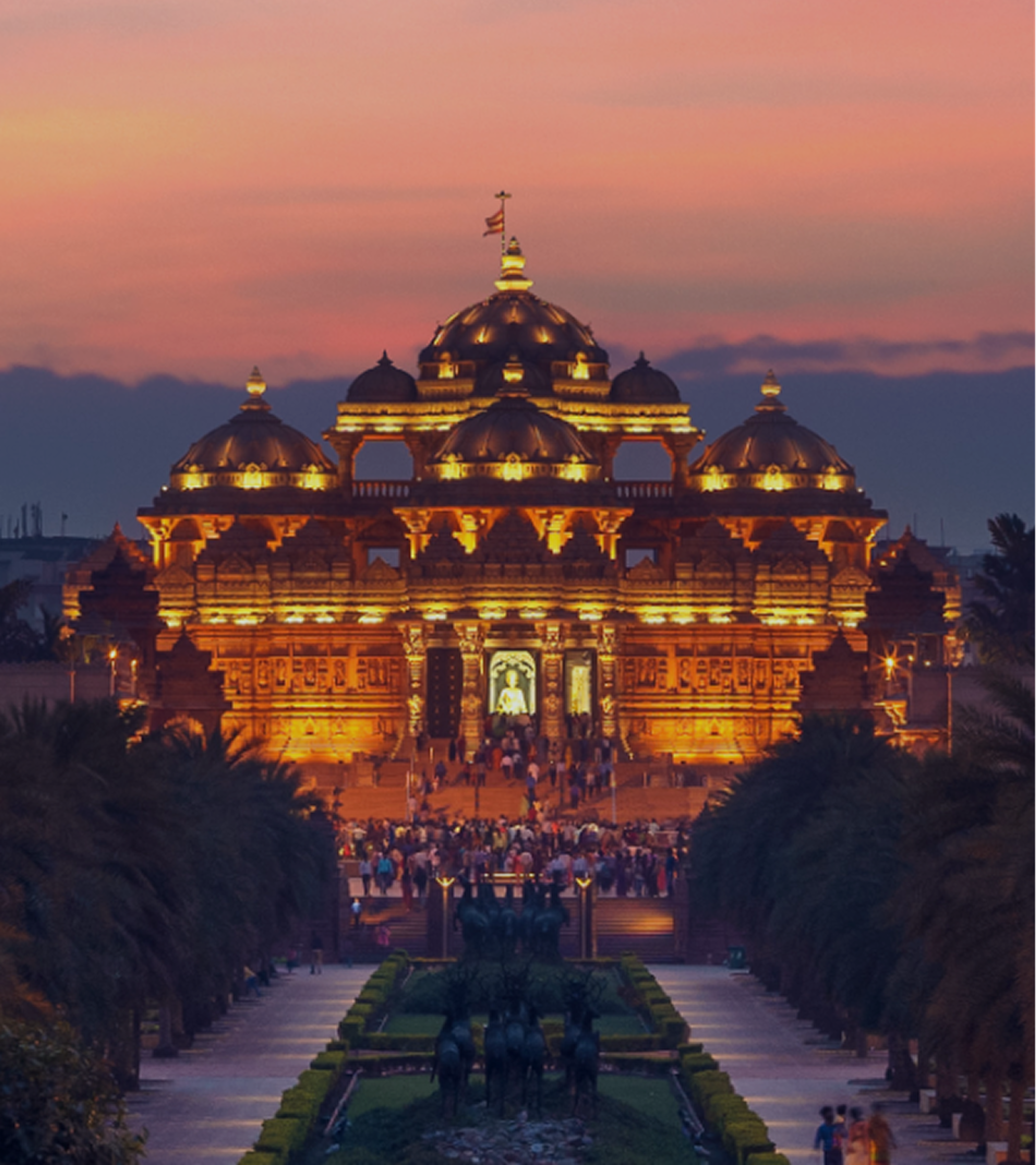 Illuminated Indian temple at sunset, with ornate domes and a palm-lined walkway leading to the entrance.