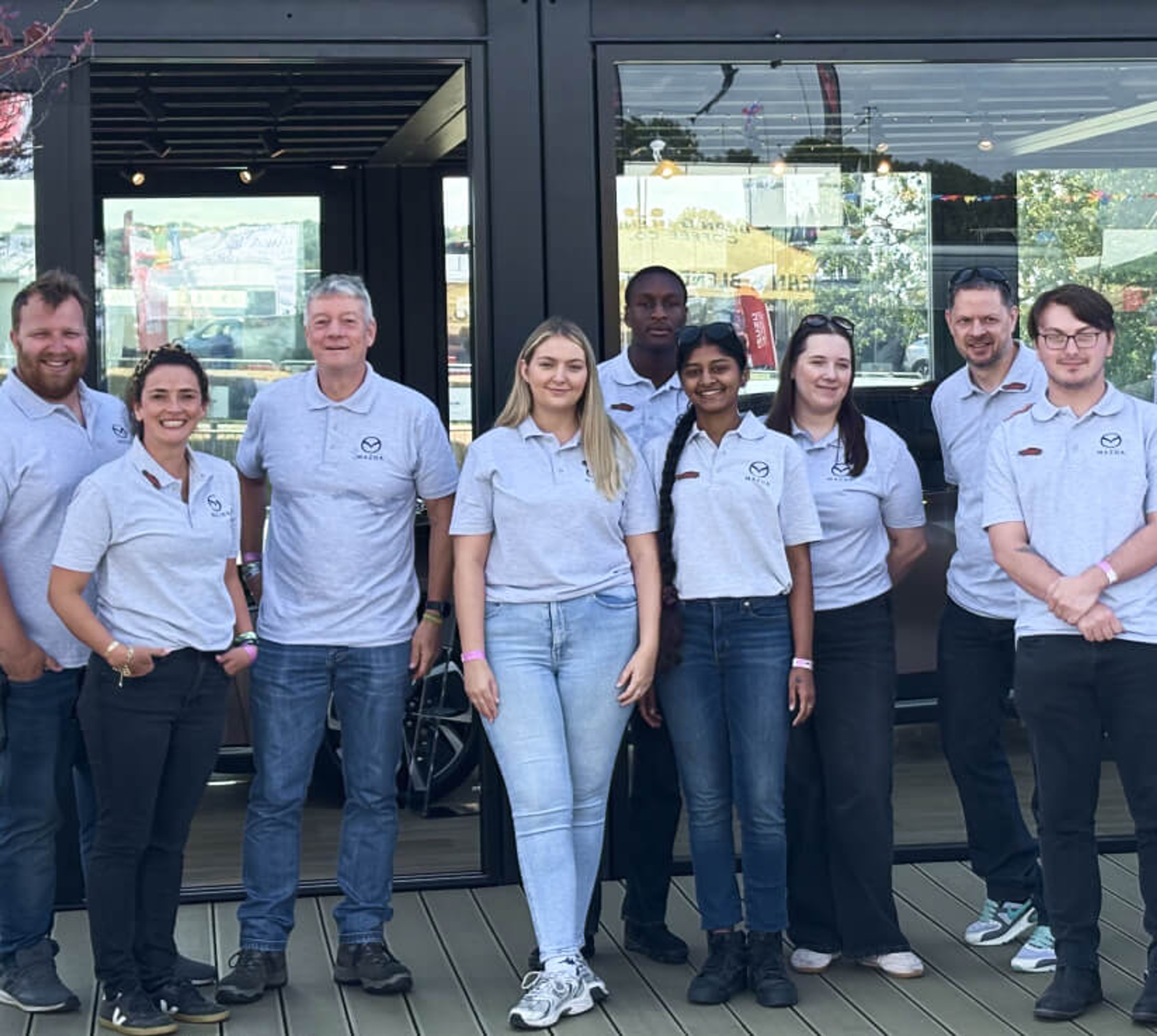 A group of corporate staff members wearing matching light grey polo shirts posing for a group photo outside a glass-fronted venue