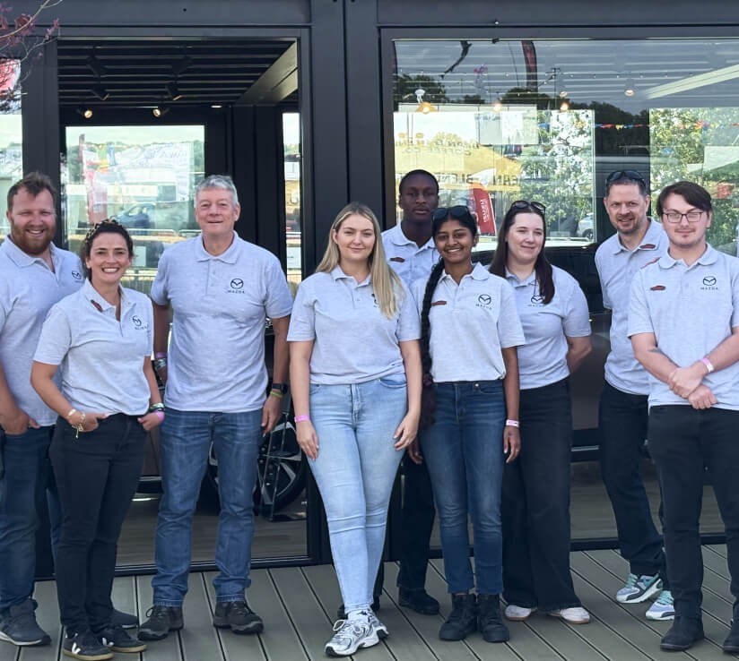 A group of corporate staff members wearing matching light grey polo shirts posing for a group photo outside a glass-fronted venue