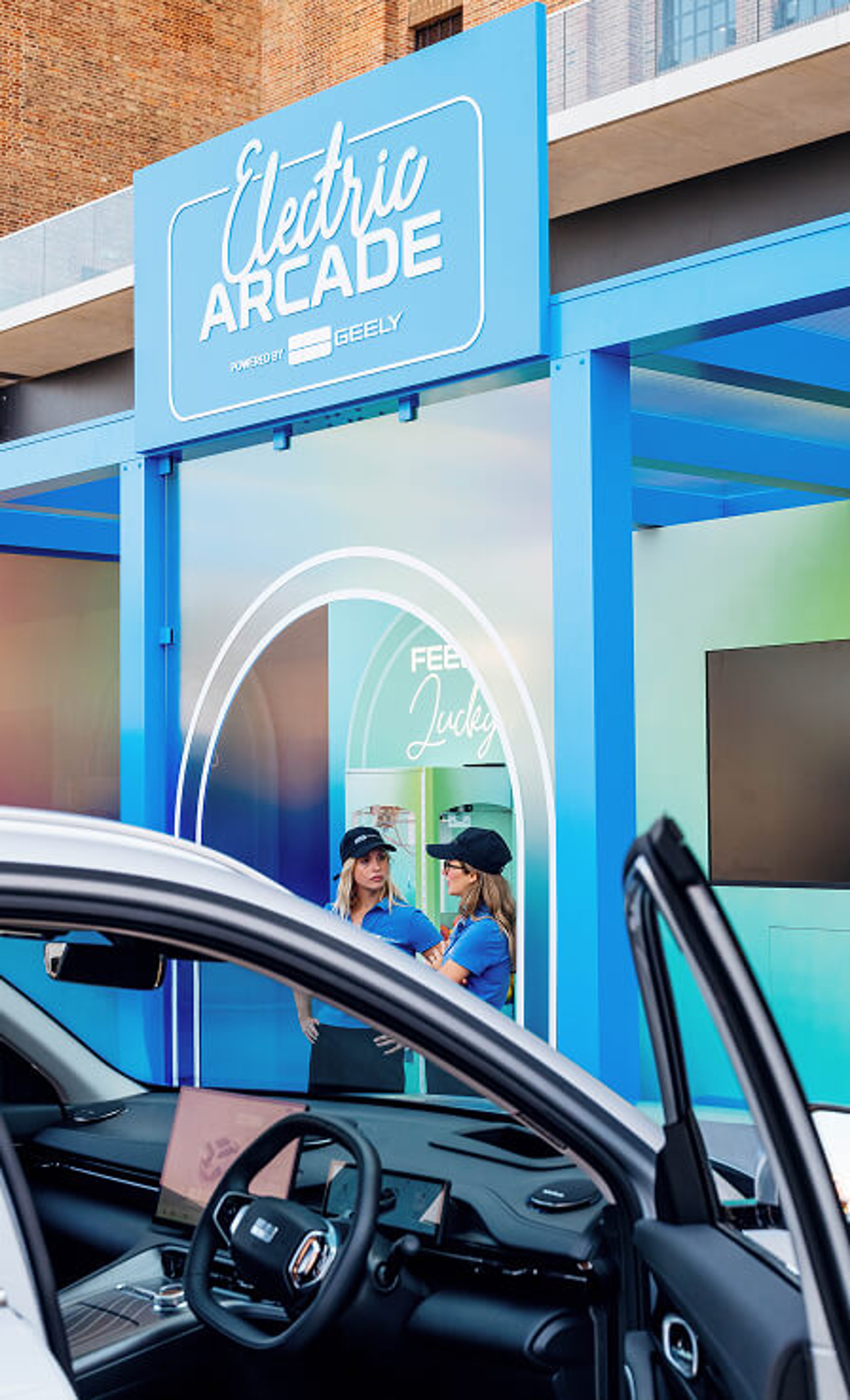 The interior dashboard and steering wheel of a car with two event staff members visible in front of an Electric Arcade