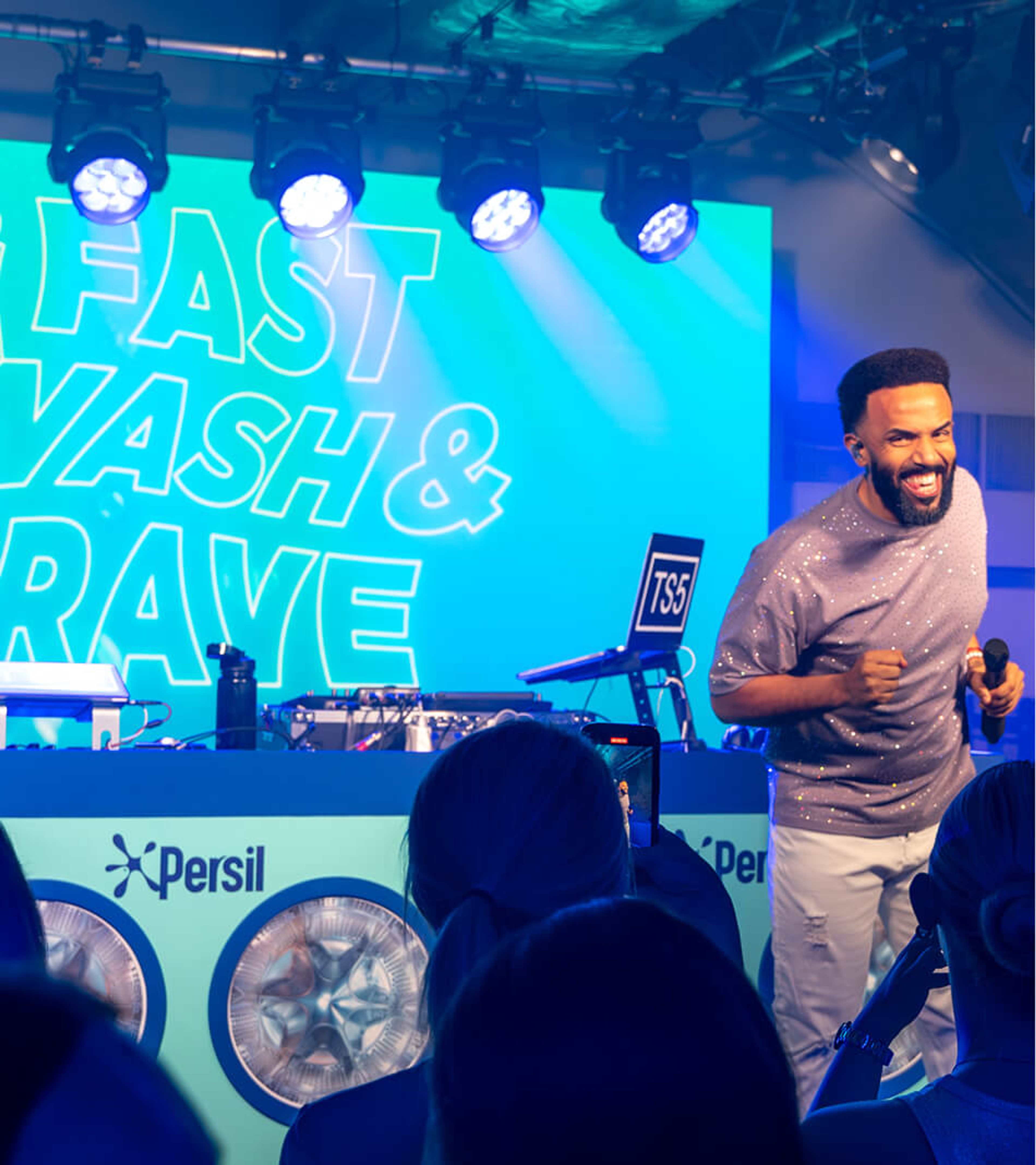 Craig David performing on stage holding a microphone in front of a blue screen reading 'Fast Wash & Rave' at a Persil event