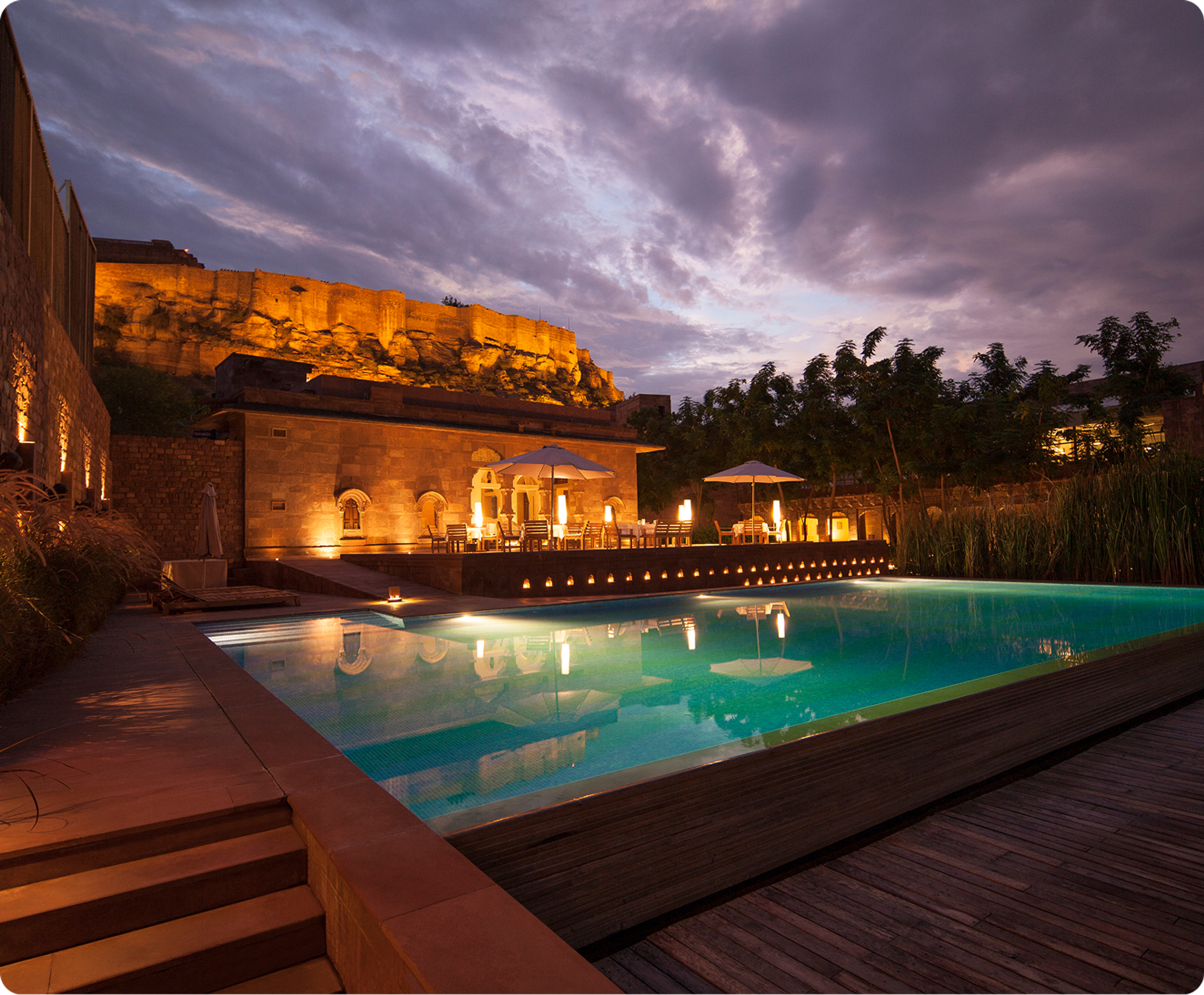 Illuminated hotel pool and terrace beneath a hilltop fort at dusk.