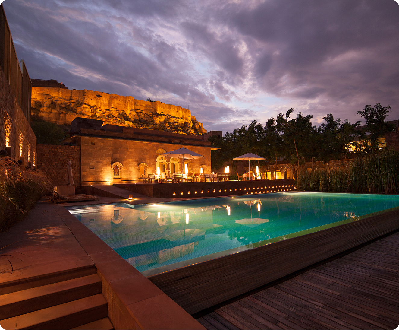 Illuminated hotel pool and terrace beneath a hilltop fort at dusk.