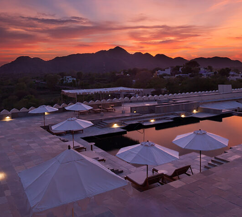 Hotel pool terrace with white parasols beneath a pink sunset and mountain skyline.