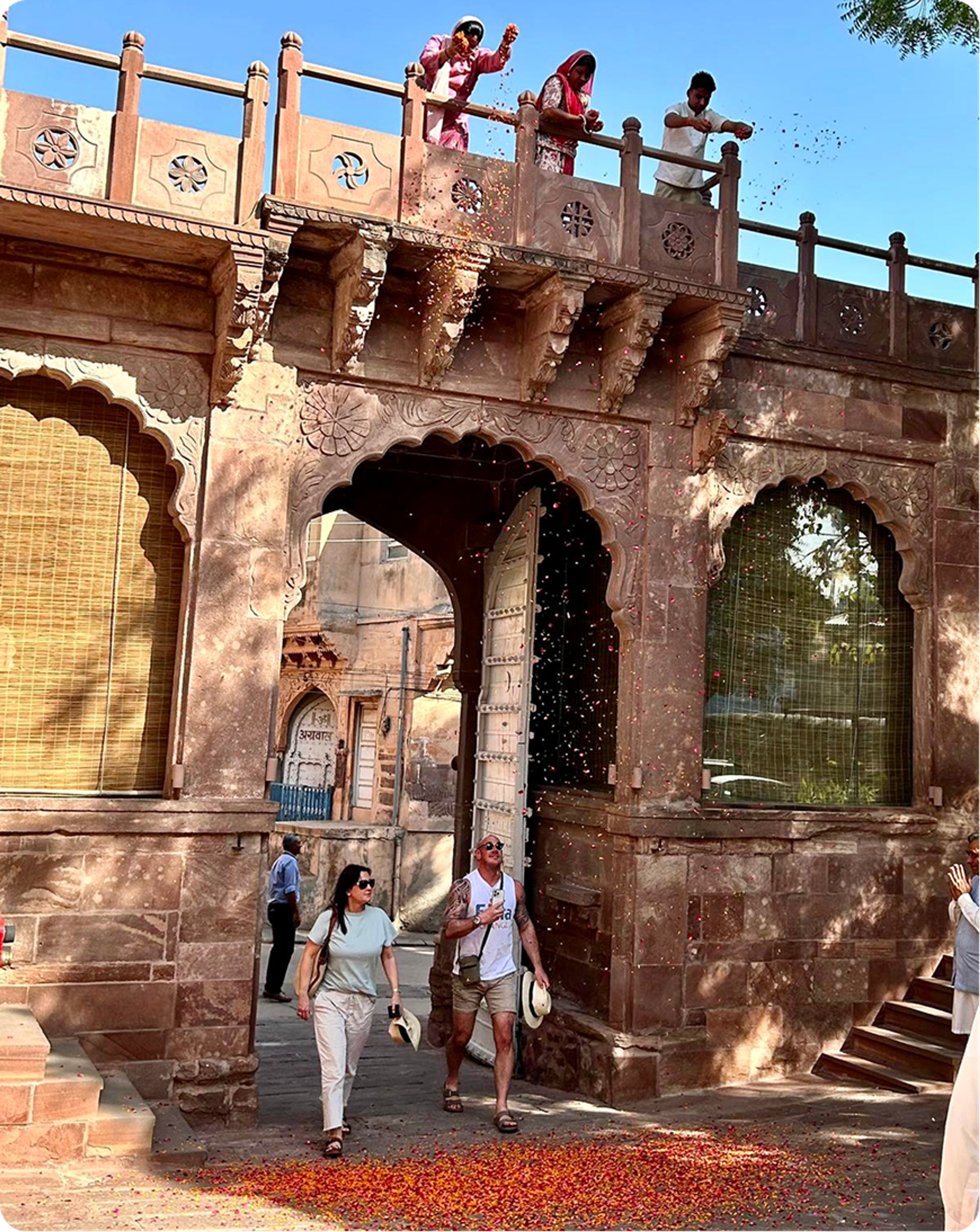 Guests walking beneath an ornate stone archway as flower petals fall around them.