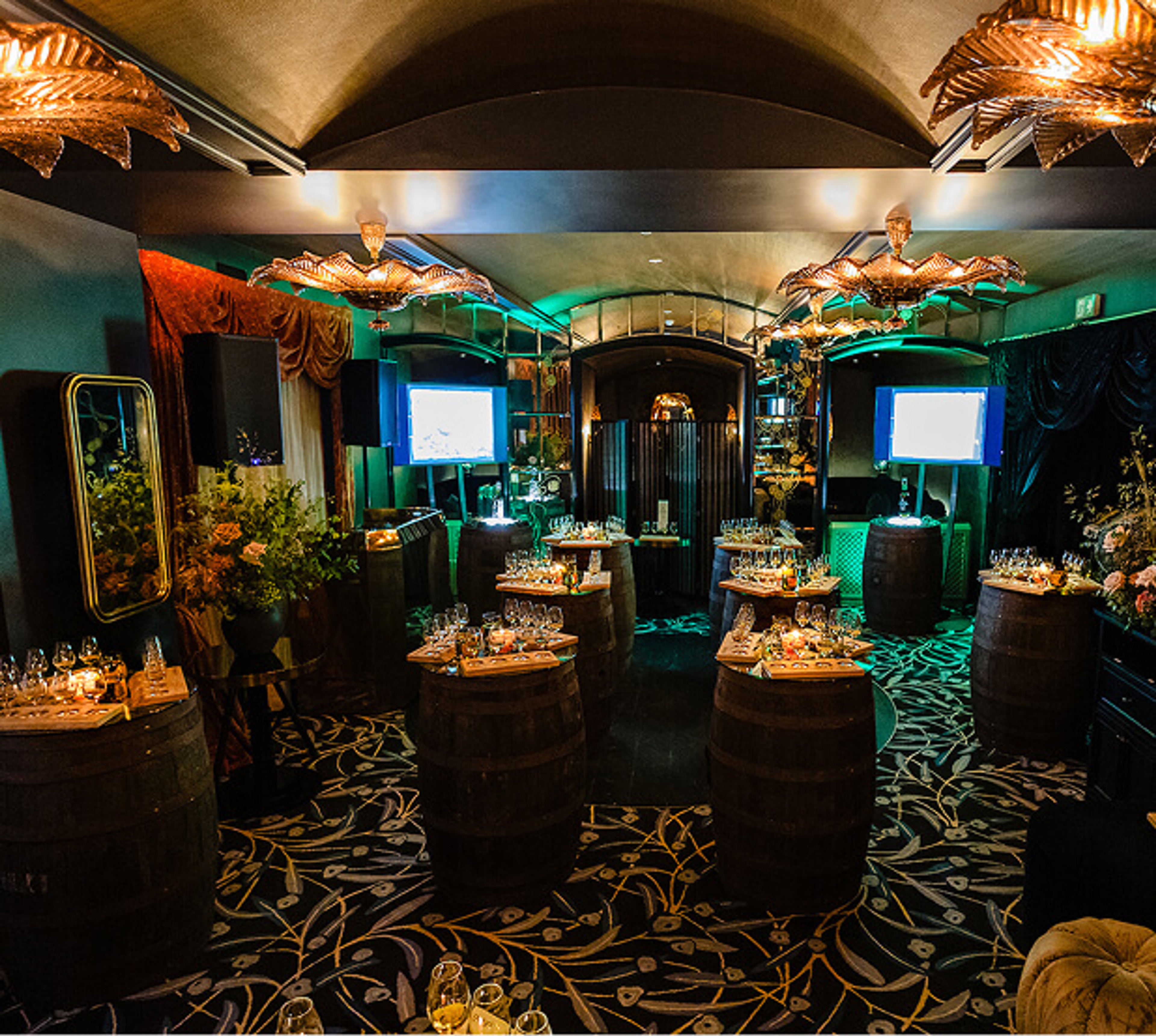Dimly lit luxury event venue decorated with wooden whiskey barrel tables, ornamental chandeliers, and ambient green lighting