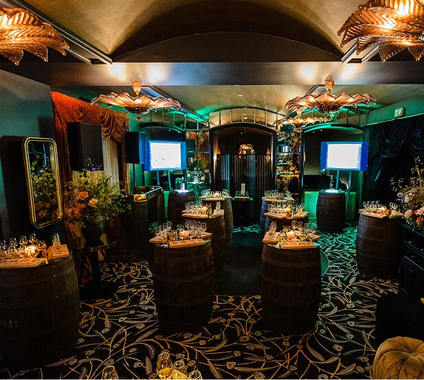 Dimly lit luxury event venue decorated with wooden whiskey barrel tables, ornamental chandeliers, and ambient green lighting
