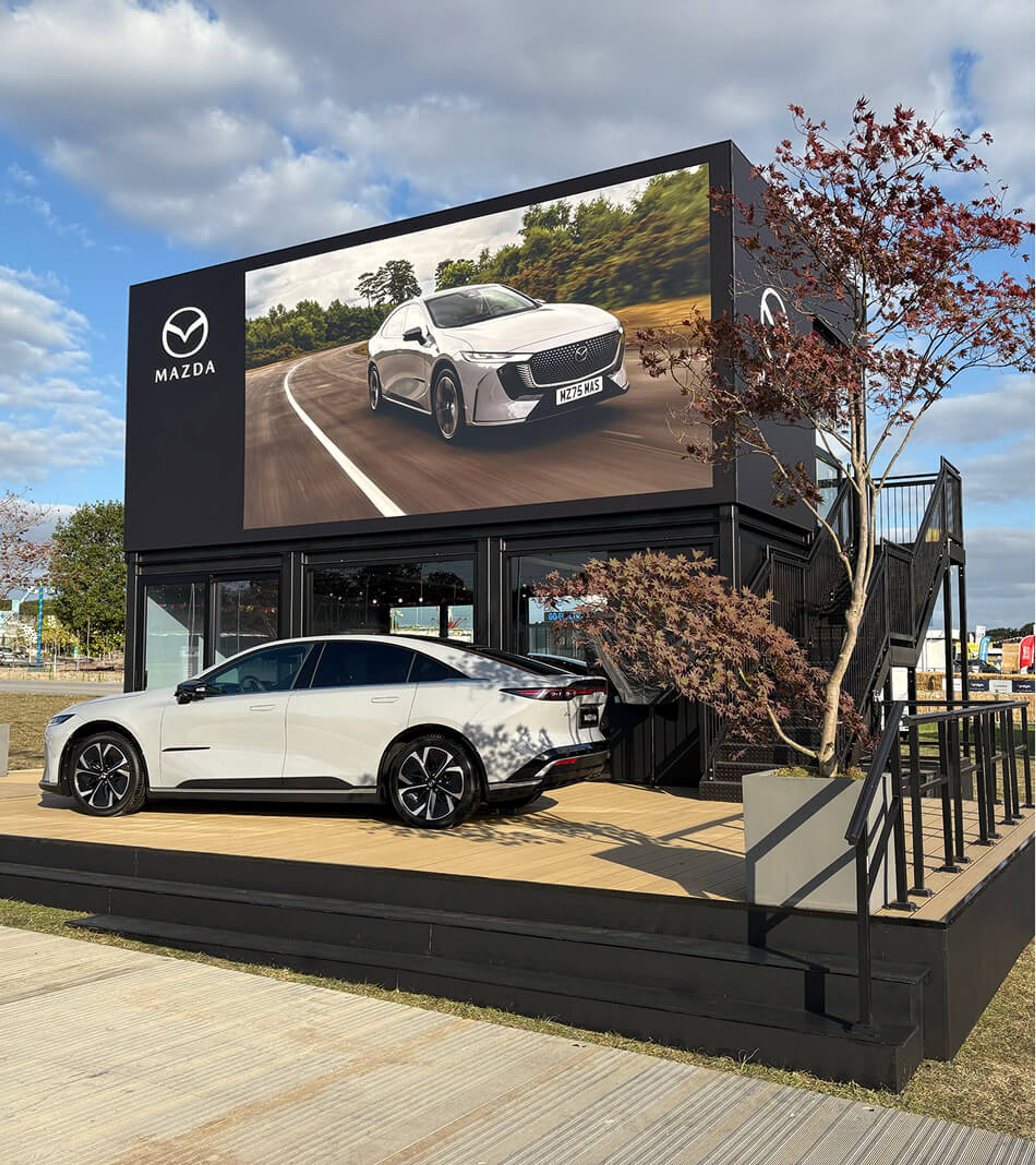 Mazda outdoor event space featuring a branded two-storey structure with a large digital screen, a Mazda vehicle on display and landscaped surroundings.