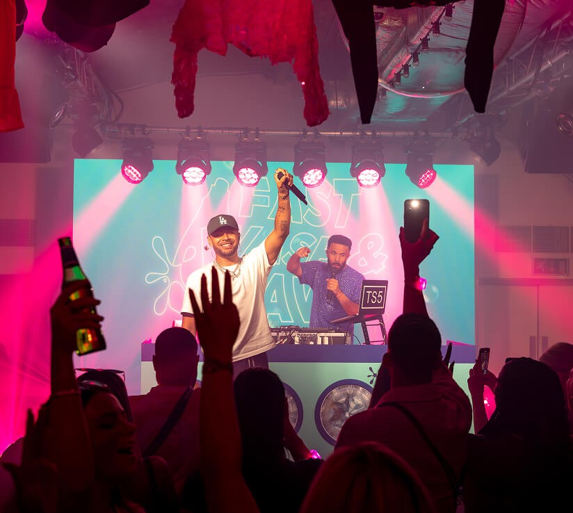 Performers on stage interacting with an enthusiastic crowd during a live music event under pink lighting