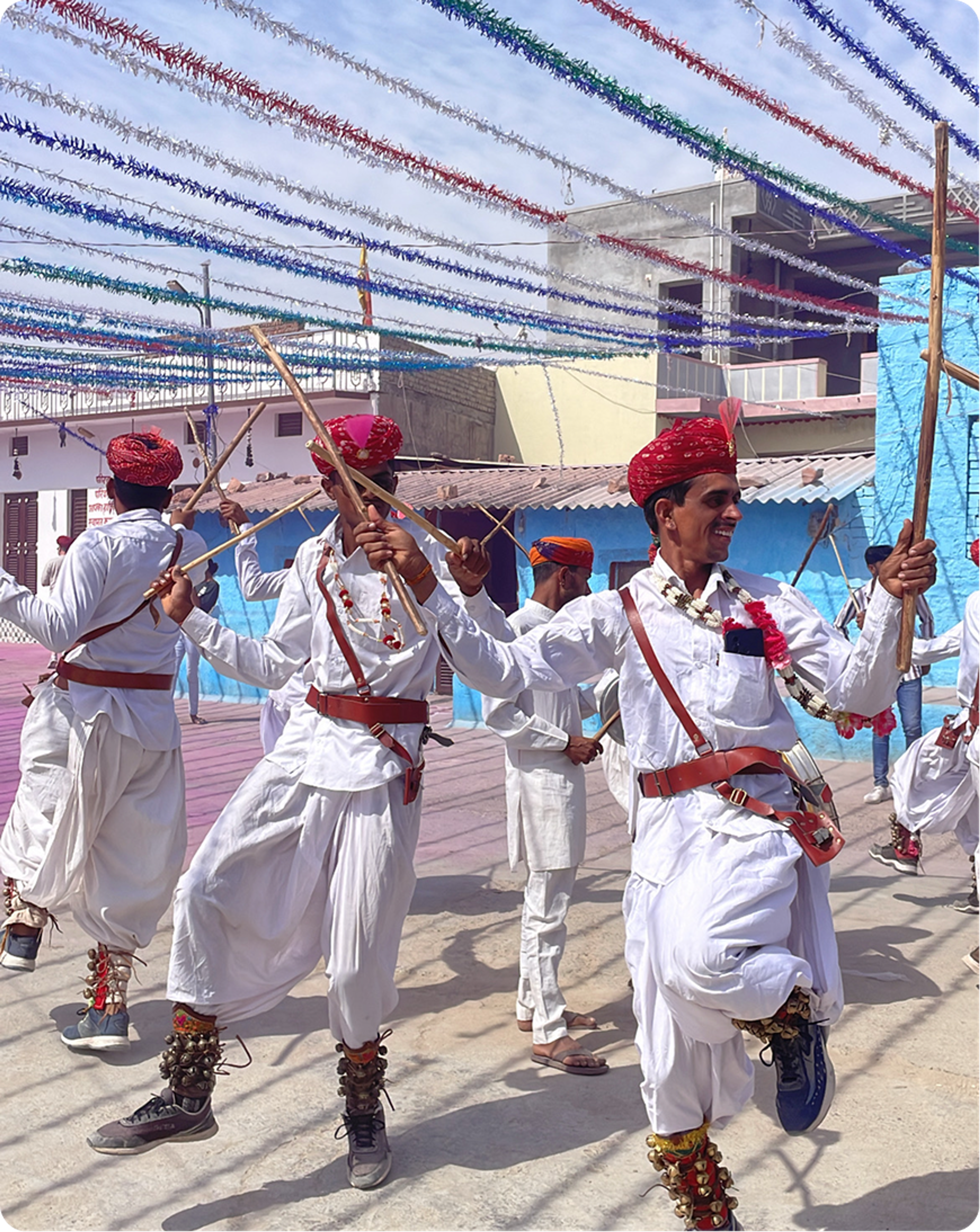 Traditional dancers in white outfits and red turbans performing with wooden sticks.