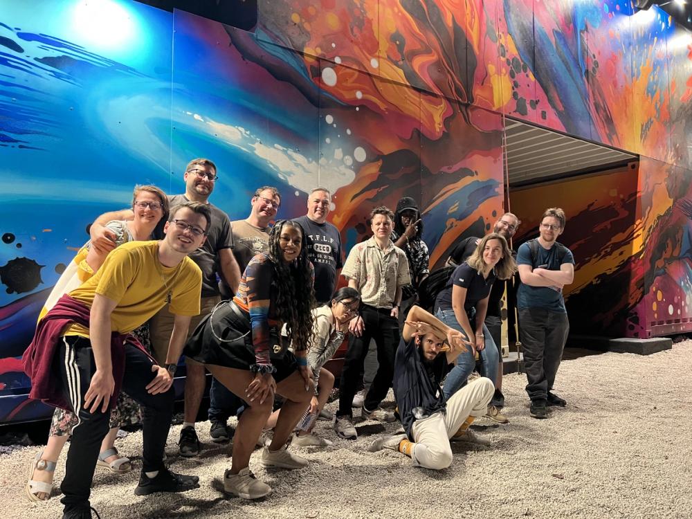 A group of thirteen people are posed in a line. The line is angled away from the photo, and runs parallel to a psychedelic art piece that runs for the entire background. Everyone in the group is smiling widely and bunched together in camaraderie.