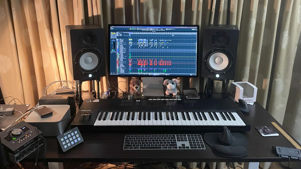 A large keyboard sits on a desk underneath a computer monitor. Either side of the monitor is a pair of large audio speakers. Various mixing and audio equipment surrounds the desk, and two small plush toys, a beaver and a mouse, watch the process!