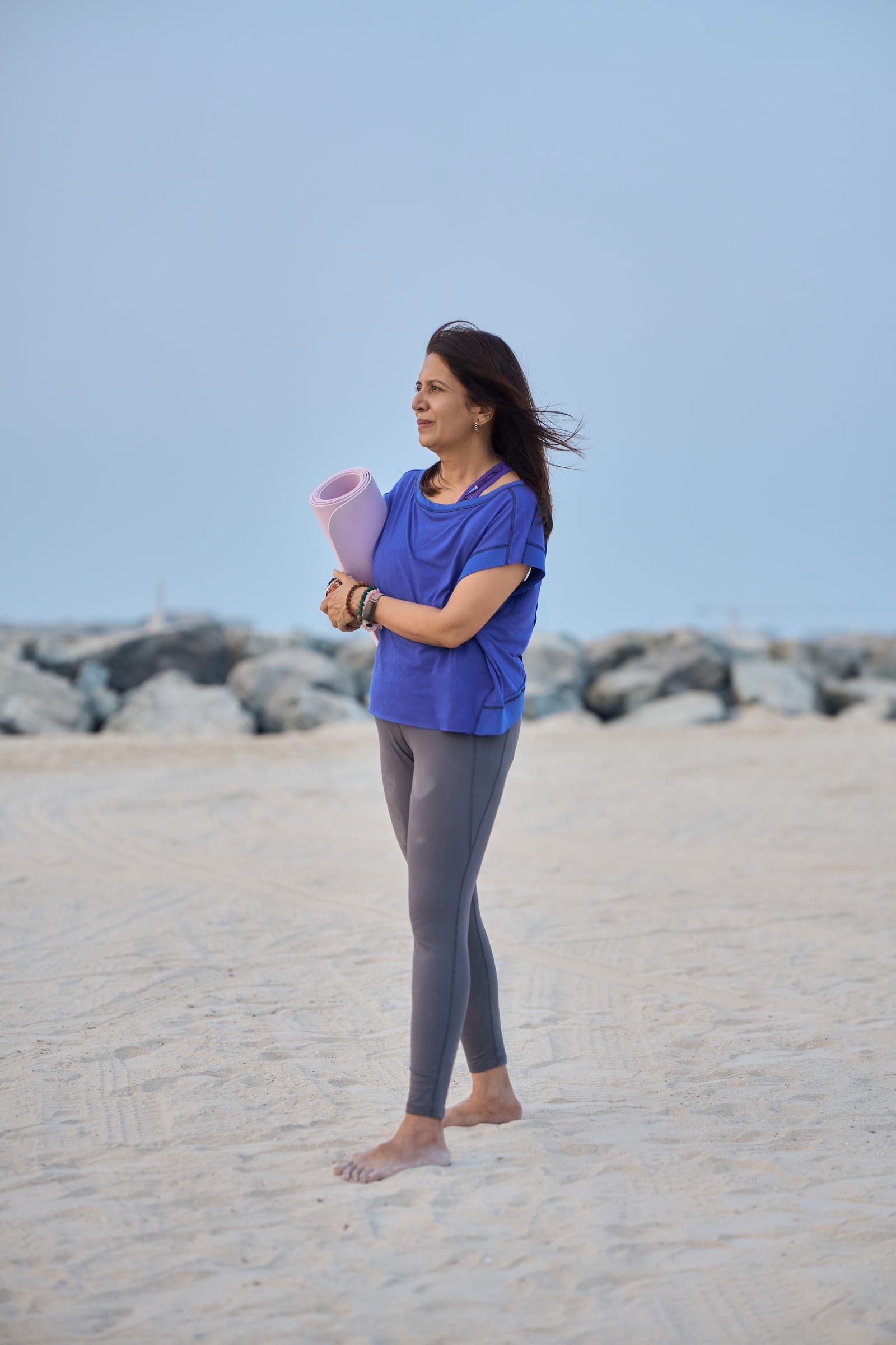Manisha standing on the beach holding her rolled yoga mat