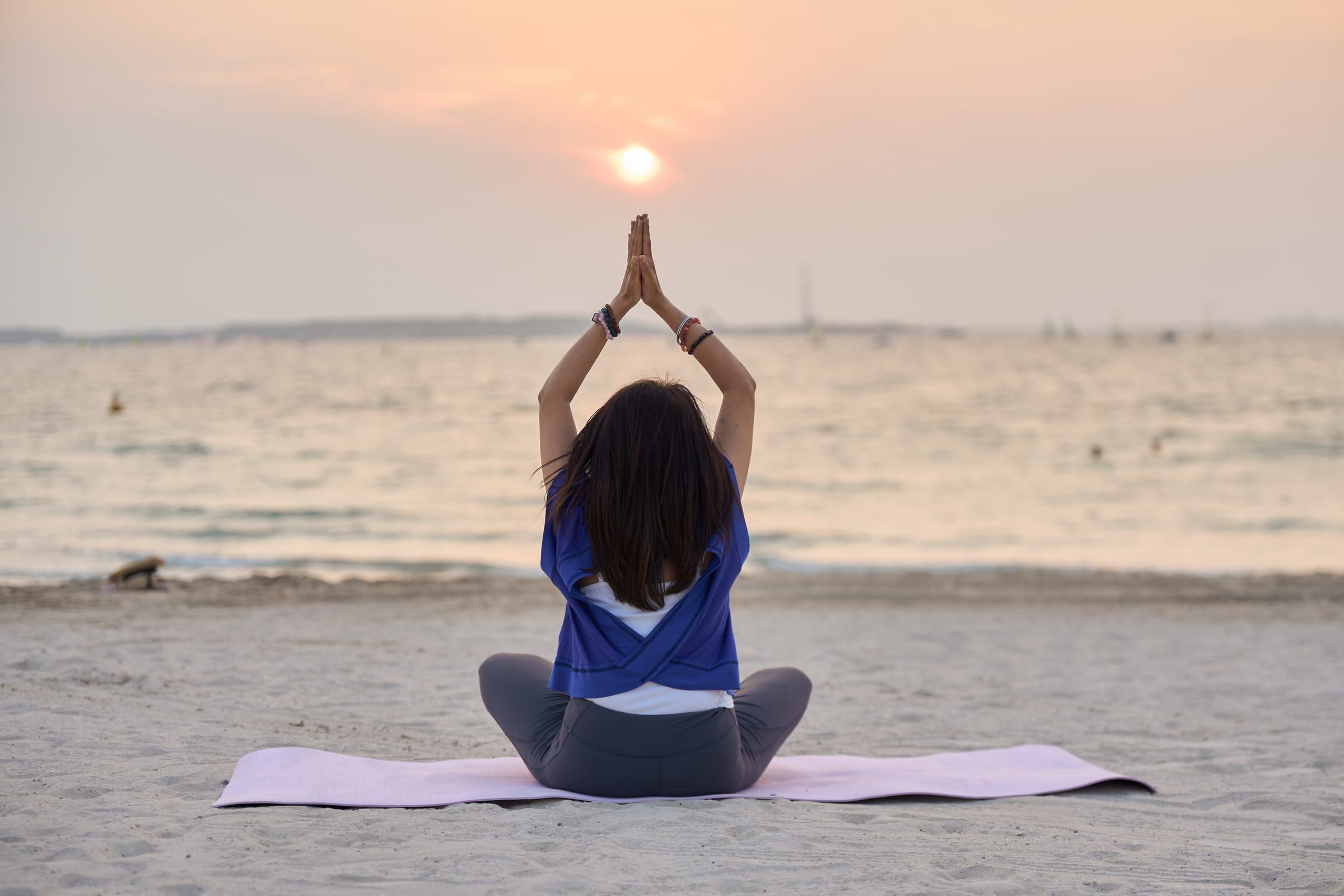 Manisha seated on a yoga mat on the beach, facing the sunset with arms raised